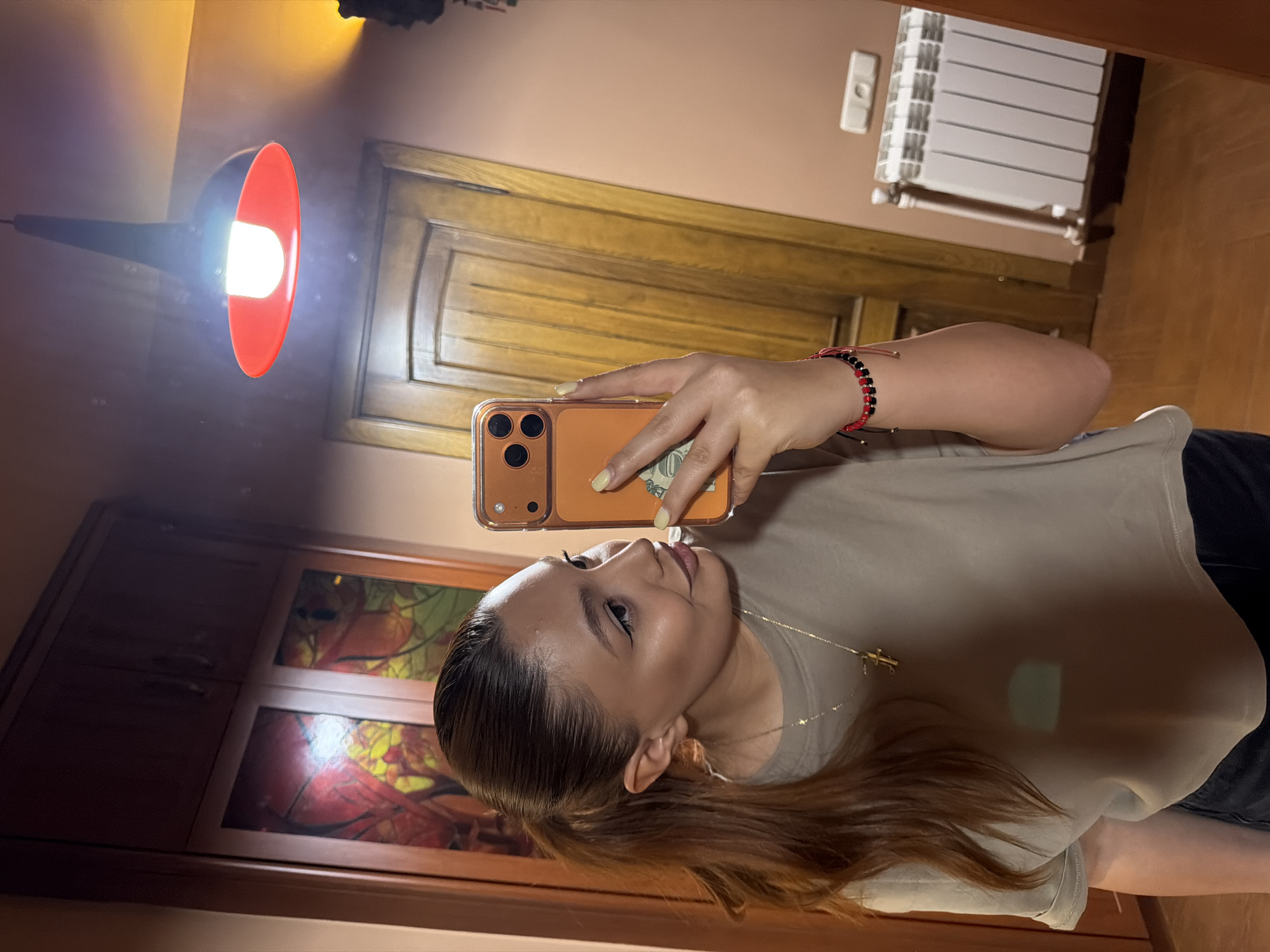A woman is taking a selfie in a room that appears to be in a residence. She is holding an orange iPhone, which obscures part of her face. Her hair is brown and pulled back in a ponytail. She is wearing a light brown t-shirt and a gold necklace with a cross pendant. She has a red and black beaded bracelet on her wrist.
Behind her, a wooden door is visible, as well as a radiator. A lamp with a red shade is positioned above and to the left, with its light source clearly visible. There is also a wooden cabinet or window frame with what appears to be a stained-glass-like design. The room has a warm-toned color scheme.
The exact location within Yerevan, Armenia, cannot be determined from this image alone. Further information or context would be required to pinpoint the specific location.