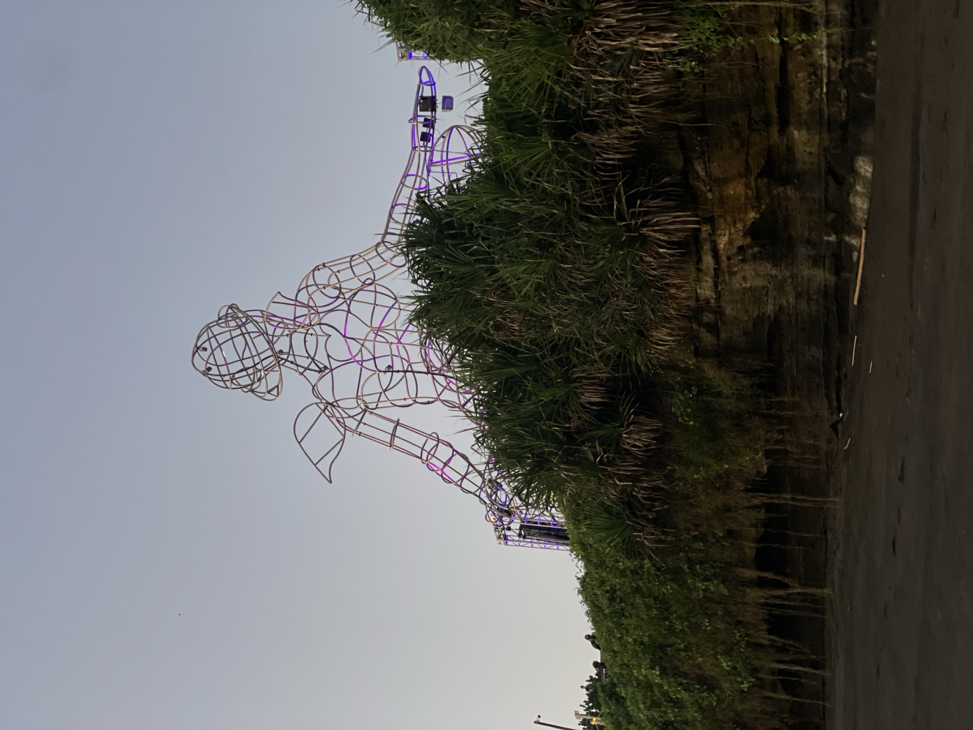 The image captures a wireframe sculpture of a human figure, illuminated by purple lights, situated atop a lush, green embankment. The sculpture is a skeletal representation, with its form defined by interconnected wires. It appears to be a large-scale installation, possibly for an event or artistic display. Beneath the embankment, a dark, sandy beach is visible, with the low tide revealing exposed rocks and hanging roots from the vegetation above. The sky is a soft, pale gray, indicating either dawn or dusk. In the background, a stage structure with more lights can be seen, suggesting the sculpture is part of a larger festival or performance area. Several figures are visible near the top of the embankment, standing around the sculpture and on the stage, though their identities are not discernible. The overall scene evokes a sense of artistic display and outdoor gathering in a coastal environment. The location is not explicitly mentioned as Kediri, Indonesia, but the description focuses on the visual elements of the image.