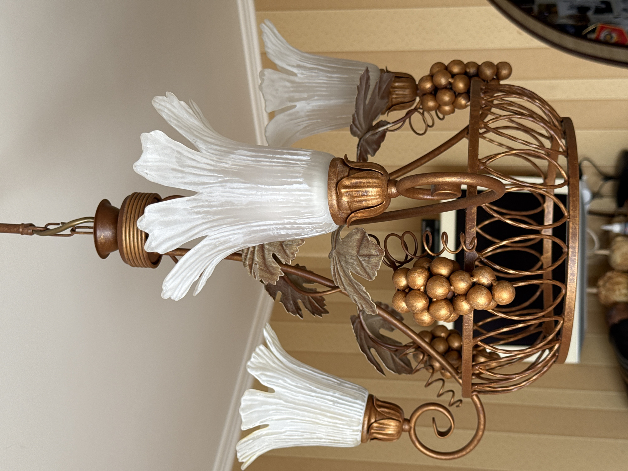 A close-up, low-angle shot captures an ornate, vintage-style chandelier hanging from a textured ceiling. The chandelier is made of a coppery-bronze metal and features three tulip-shaped frosted glass shades, each with delicate vertical ridges. Decorative elements include stylized grapevine leaves and clusters of faux golden grapes adorning the fixture. The background consists of a wall with vertical yellow stripes.