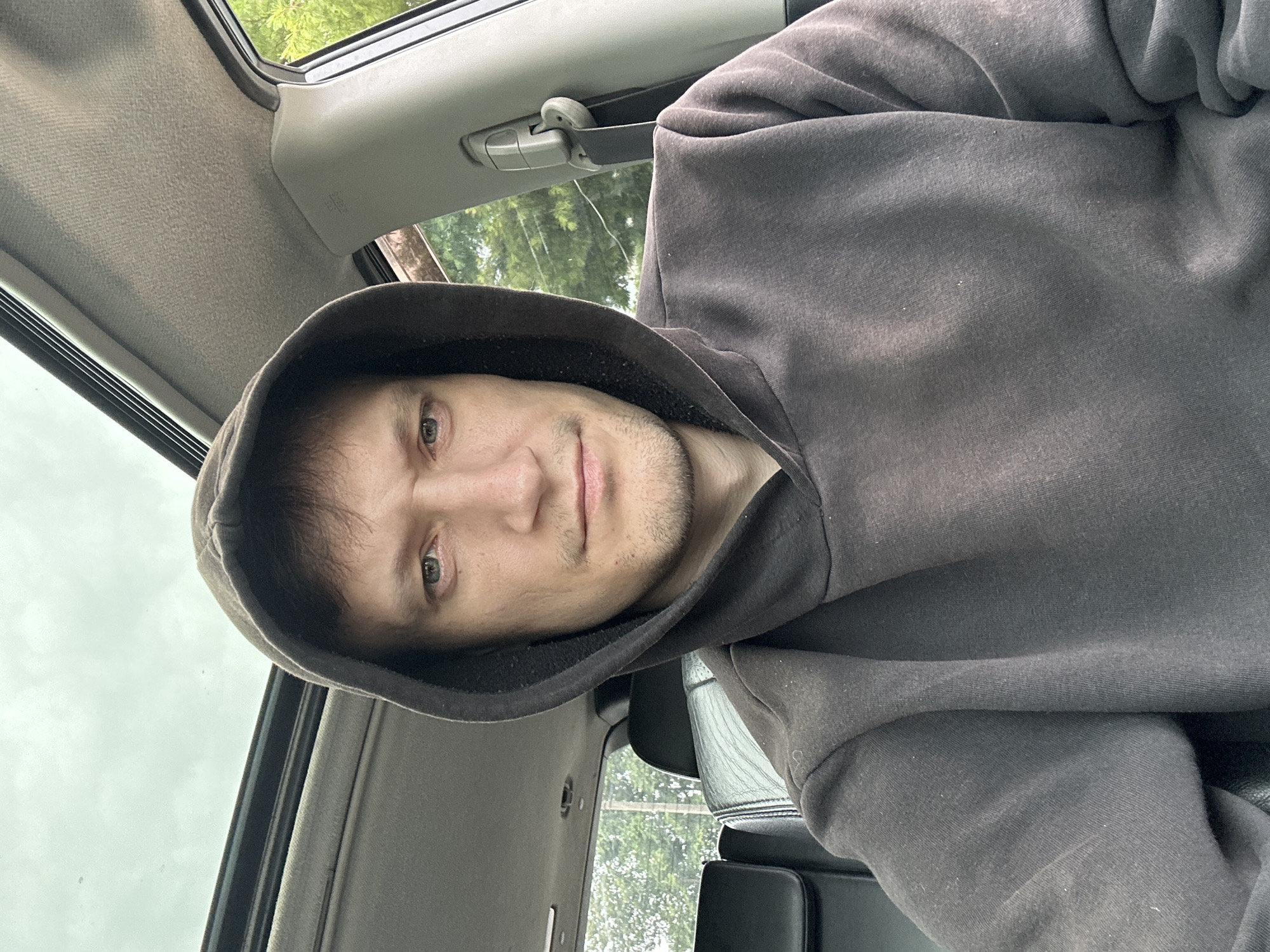 A man wearing a dark gray hooded sweatshirt is seated inside a vehicle, looking directly at the camera. He has light skin, dark hair, and a neutral expression with his mouth slightly downturned. The vehicle's sunroof is open, revealing a cloudy sky above. The interior of the car shows a gray headliner and part of a black seat. Green foliage is visible through the open window. The man's hands are not visible. The image appears to be a selfie taken from the driver's seat. The location is not identifiable as Makiv, Ukraine, or any specific city.