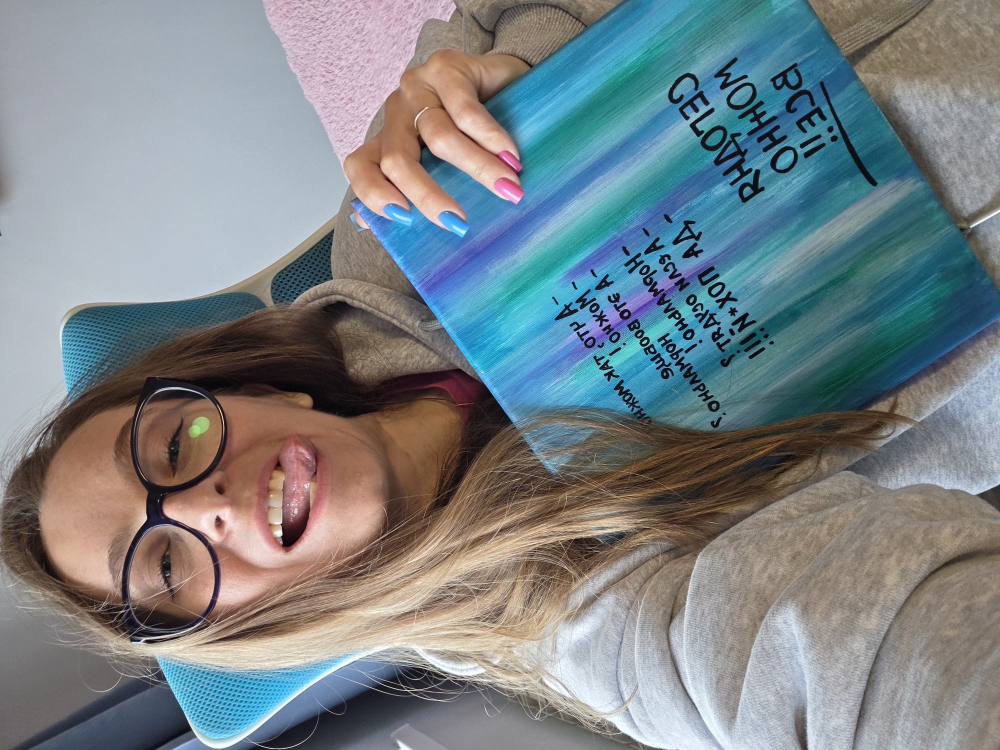 A woman with blonde hair and glasses is sitting in a chair, holding a painting. She is sticking her tongue out and making a face at the camera. The painting is abstract, with streaks of blue, green, and purple. There is text on the painting, but it is in a foreign language and difficult to read. The woman is wearing a gray hoodie. The room appears to be a bedroom or living room, with a pink blanket visible in the background. There is no visible indication of the city of Birobidzhan, Russia.