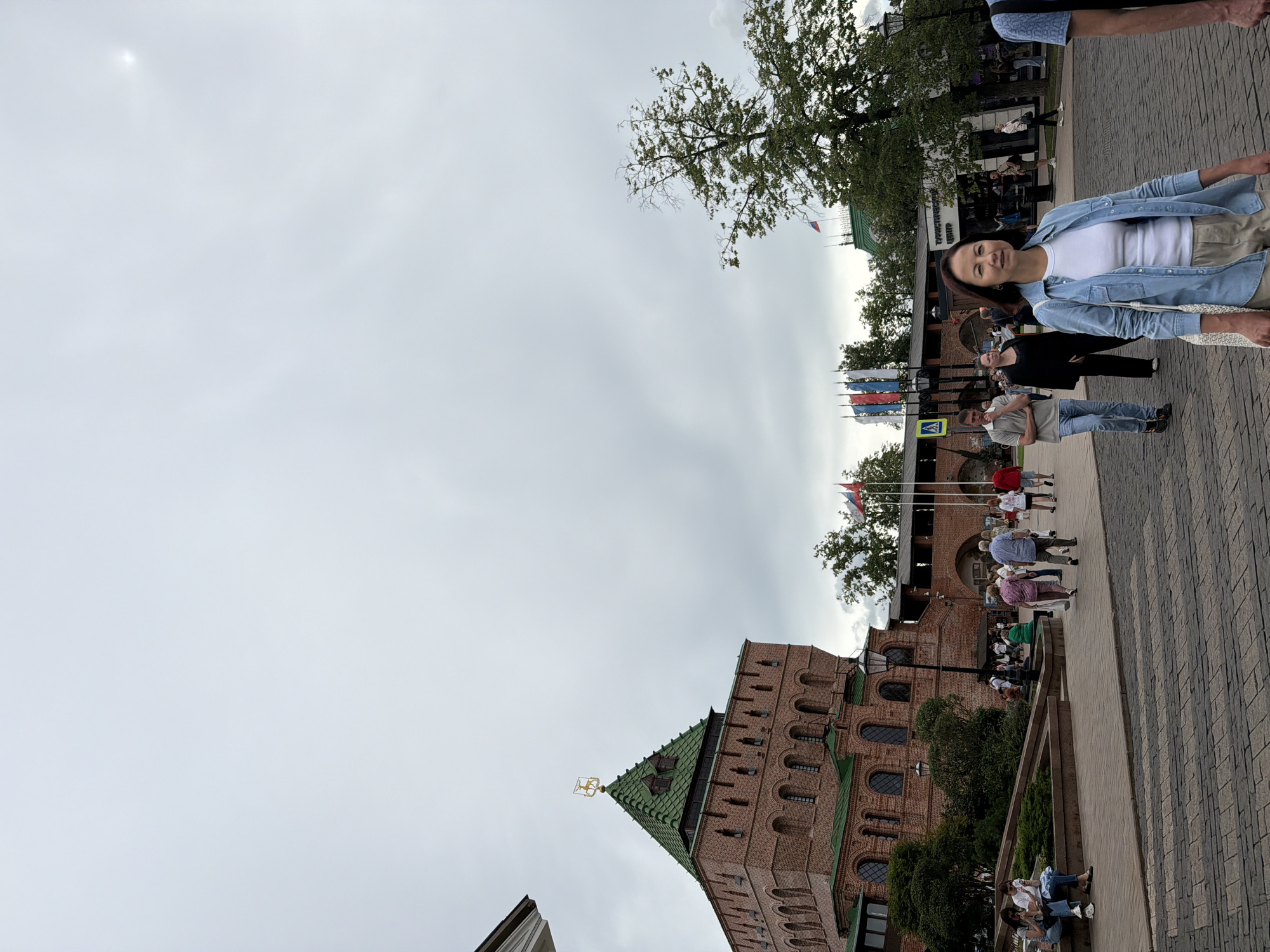 The image depicts a street scene in Nizhny Novgorod, Russia, likely at the Kremlin. In the foreground, a woman with dark hair, wearing a light blue denim jacket over a white t-shirt and beige pants, walks towards the camera. She carries a white woven bag over her right shoulder. Behind her, other pedestrians are visible. To the left, a large, red brick tower with a green roof and a golden deer statue on top is prominent. This is likely one of the towers of the Nizhny Novgorod Kremlin. The background features more brick structures, trees, and flags. The sky is overcast.
