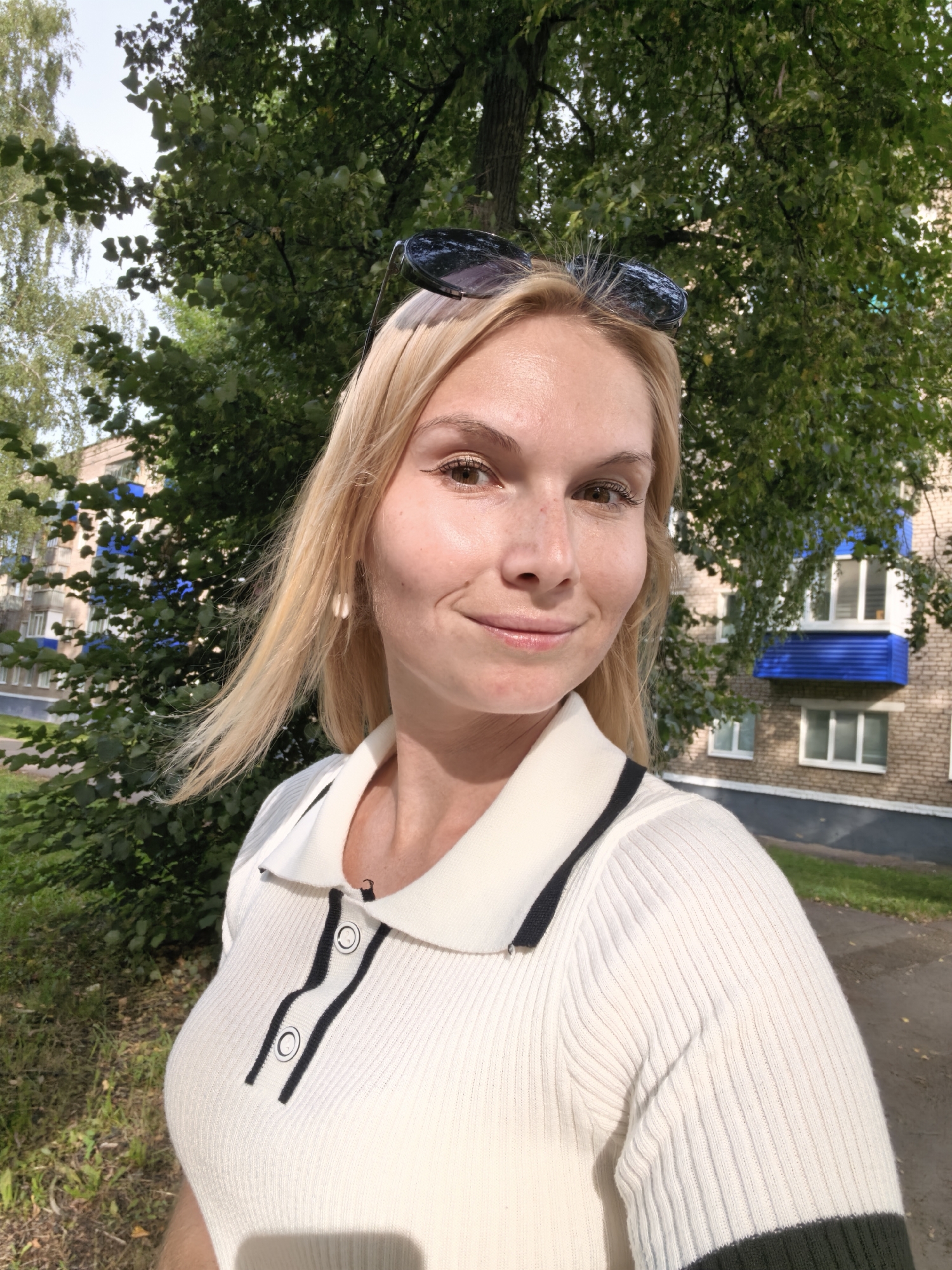 The image depicts a young, fair-skinned woman with blonde hair and light brown eyes, posing for a selfie outdoors. She is wearing a white, ribbed polo shirt with black trim and buttons. Sunglasses are perched on top of her head. The background reveals a brick apartment building with blue accents on balconies, typical of Soviet-era architecture, suggesting a location in the city of Sterlitamak, Russia. Lush green trees and foliage frame the scene, indicating an outdoor setting, possibly a residential area. The lighting suggests it is daytime, with sunlight casting shadows and highlighting the woman's features.