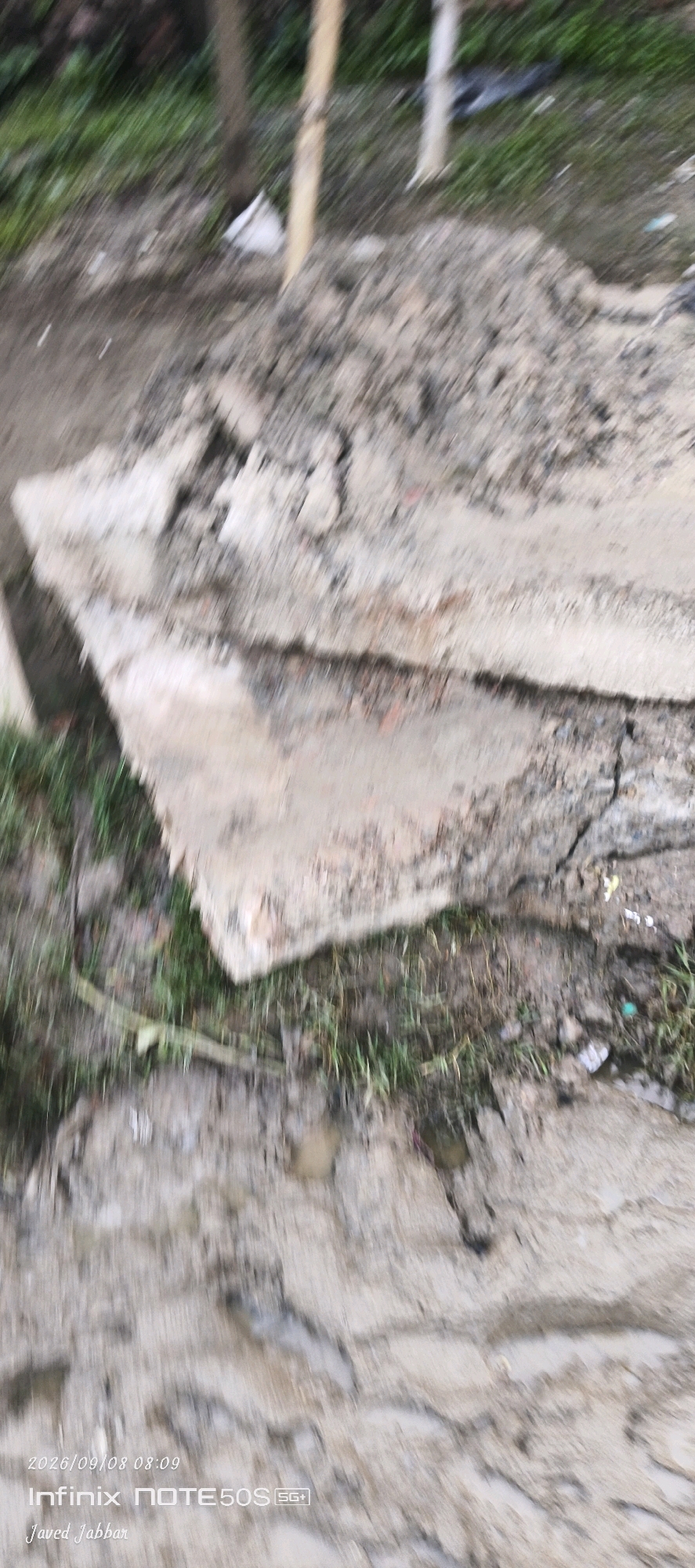 The image shows a close-up, blurry view of a construction or demolition site. Large pieces of concrete or rubble are scattered on the ground, mixed with dirt and grass. A thin wooden or bamboo pole is visible vertically in the background, along with some green vegetation. The overall impression is of an unkempt outdoor area. There are no people or animals visible in the image. The text at the bottom indicates the date and time as 2026/09/08 08:09, and mentions "Infinix NOTE50S 5G+" and "Javed Jabbar". The location is stated as Bahera, India.