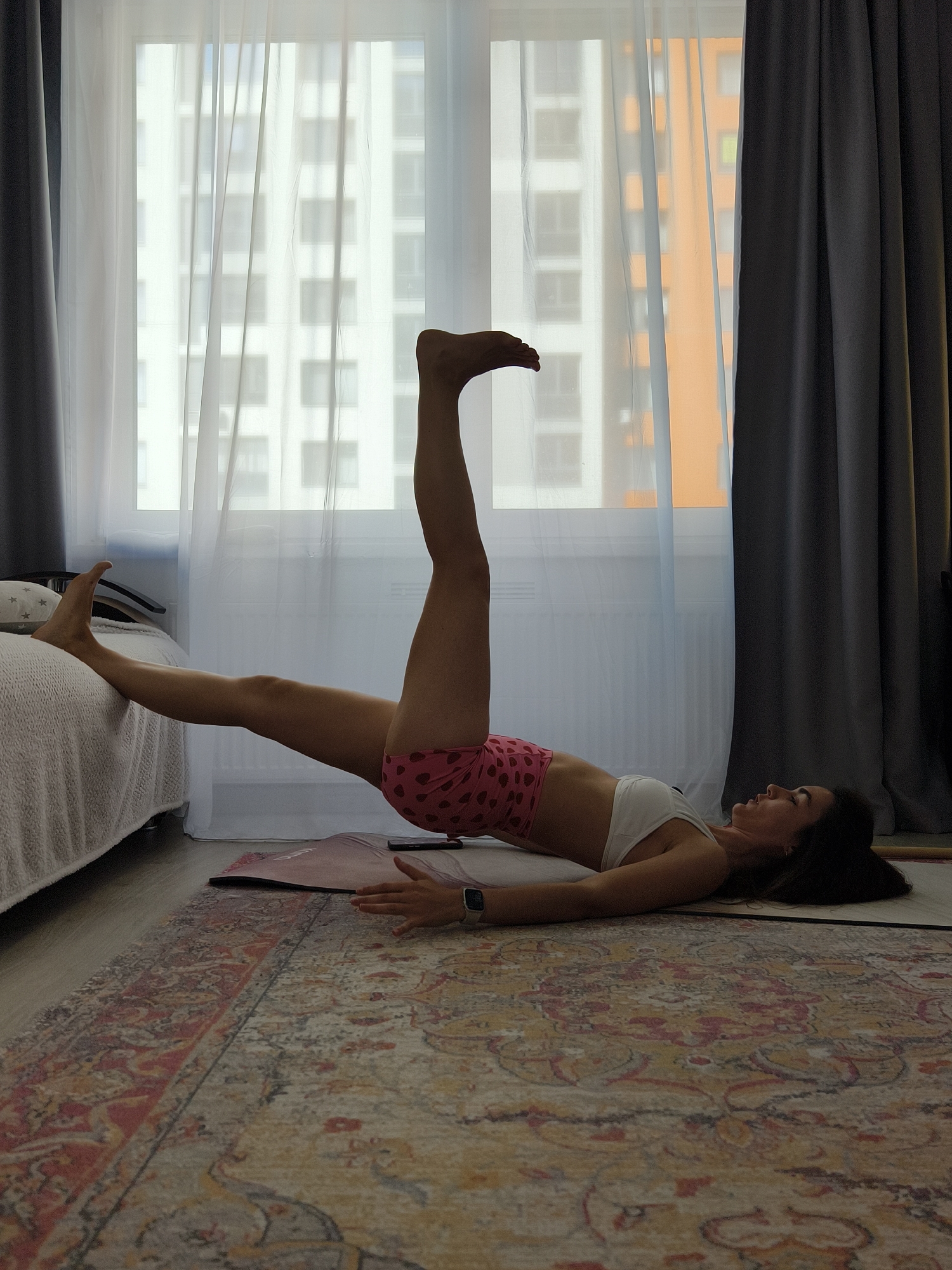 A woman lies on a yoga mat on a patterned rug, with one leg extended vertically in the air and the other resting on a bed. She wears pink shorts with a pattern and a white sports bra. The scene is indoors, likely a home setting with a window providing natural light. The window reveals a view of high-rise buildings, indicating an urban area. Curtains frame the window on either side. The city is Tyumen, Russia, as specified.