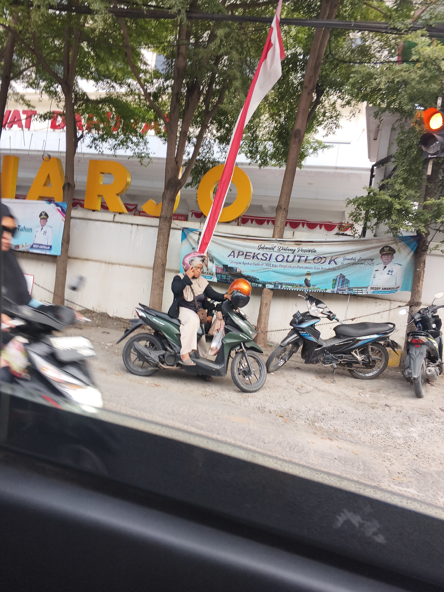 In Bandar Lampung, Indonesia, a woman wearing a headscarf and a pink helmet rides a green motorcycle. She is looking down and talking on her cell phone. Behind her, a black motorcycle is parked. Further back, another black motorcycle is visible. In the background, a banner displays "Selamat Datang Peserta APEKSI OUTLOOK Bandar Lampung 19-20 September 2023" and a portrait of DEDDY AMARULLAH. Large yellow letters, possibly part of a building sign, are visible above the banner. A red and white Indonesian flag is also prominent in the image. Trees with green leaves line the street. A traffic light, with its orange light illuminated, is visible on the right side of the frame. The foreground shows a dark, reflective surface, possibly a car window.