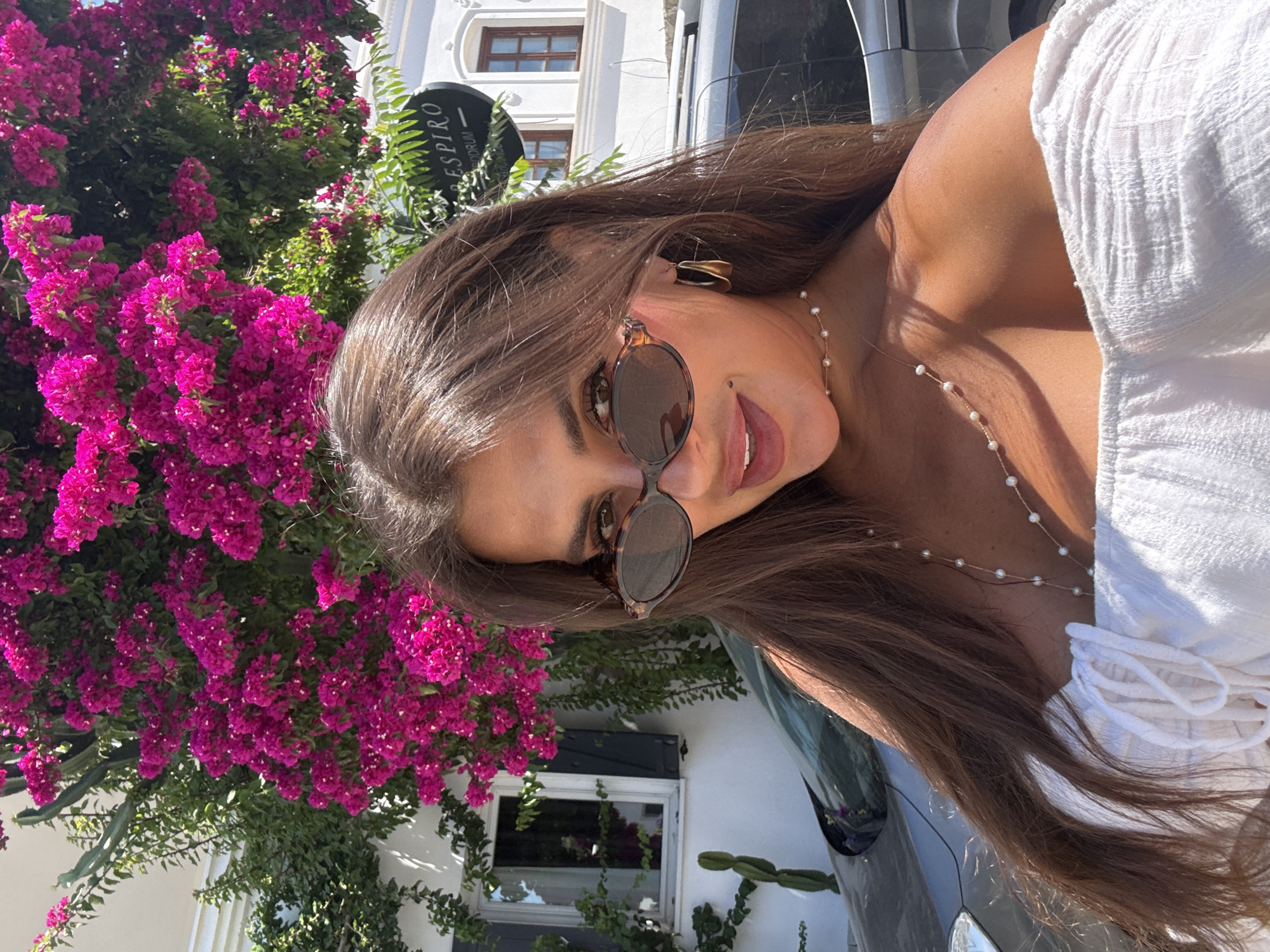 A young woman with long, brown hair and sunglasses is posing for a selfie outdoors in Bodrum, Turkey. She is wearing a white, off-the-shoulder top and a pearl necklace. Behind her, a building with bougainvillea flowers and a sign that reads "ESPIRO BODRUM" is visible. A car is parked in the background. The scene appears to be on a sunny day.