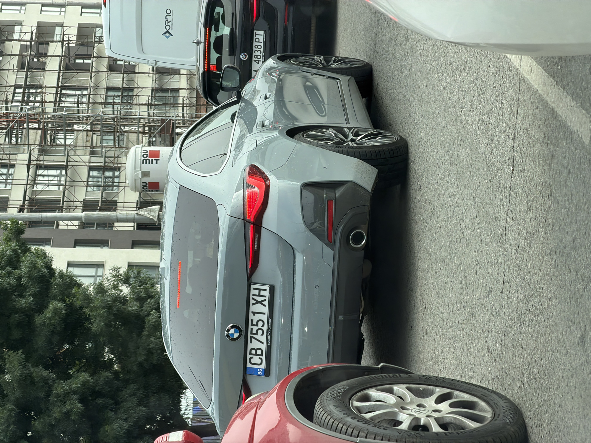 This vertical image captures a busy urban street scene in Sofia, Bulgaria, likely during the daytime under overcast or damp conditions, as indicated by the wet appearance of the vehicles and road surface.
The primary subject is a light gray BMW car, seen from the rear three-quarter view, positioned in what appears to be a line of traffic or parked cars. Its Bulgarian license plate, prominently displayed, reads "BG CB 7551 XH". Below the numbers, smaller text states "> MOBILITY.SCHWARZ". The car features multi-spoke alloy wheels and a single exhaust pipe. Water droplets are visible on its bodywork, suggesting recent rain.
In the immediate foreground, the right front wheel and a portion of a red car are visible, indicating it's directly behind the viewer's vehicle. The tire on the red car is a "Continental" brand, with "225/60 R16" partially legible.
Behind the gray BMW, a dark-colored car is partially visible, also displaying a Bulgarian license plate: "A 4838 PT". Further back and to the left, a white cargo van is seen. On its rear door, mirrored text that appears to be "ОЛГО ДЕКОР" (OLGO DEKOR) with a diamond-shaped logo is visible, likely a company advertisement.
The background features a large, multi-story building under construction or renovation, heavily covered in metal scaffolding. Below this building, two cylindrical containers or mixers are visible, one displaying the text "MIT" and the other "BAU", likely associated with construction work. To the left of the scene, lush green trees partially obscure other buildings. The road surface is asphalt, showing some cracks.