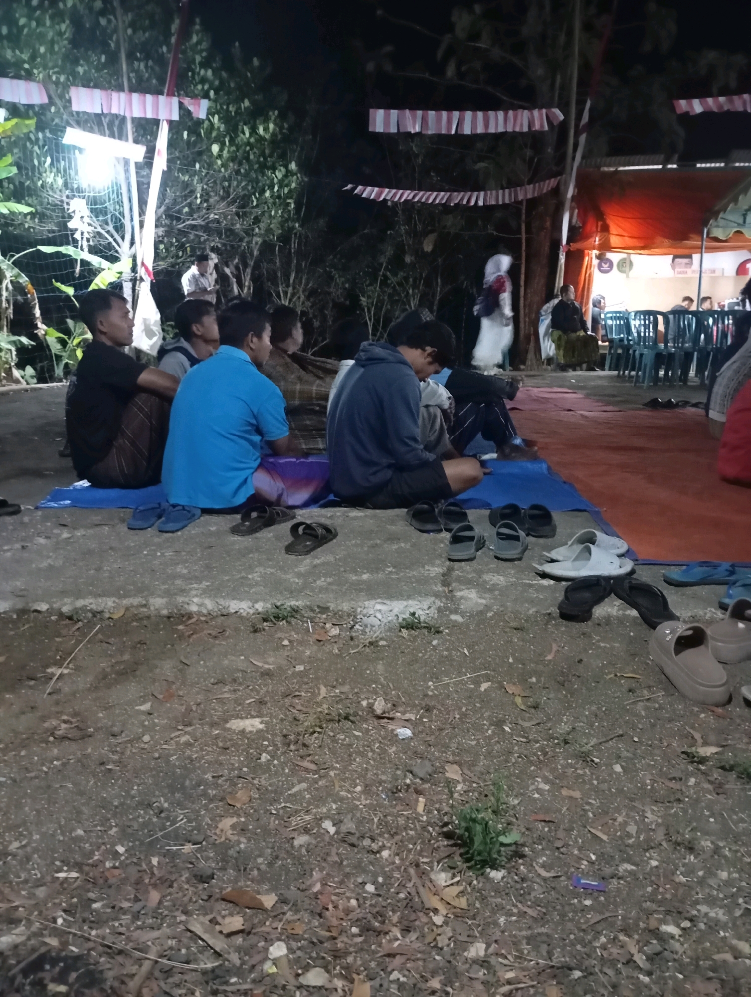 A group of people are seated on the ground in an outdoor area at night, likely a gathering or event in Wonogondo, Indonesia. The lighting is dim, with a bright light source visible in the background. Red and white banners are strung across the scene, suggesting a festive or patriotic occasion. Several individuals are facing away from the camera, some appearing to be listening or observing something. A tent structure with orange fabric is set up in the background, and rows of plastic chairs are visible under it. Various types of footwear, including flip-flops and sandals, are scattered on the ground in the foreground. The ground is a mix of dirt, gravel, and fallen leaves.