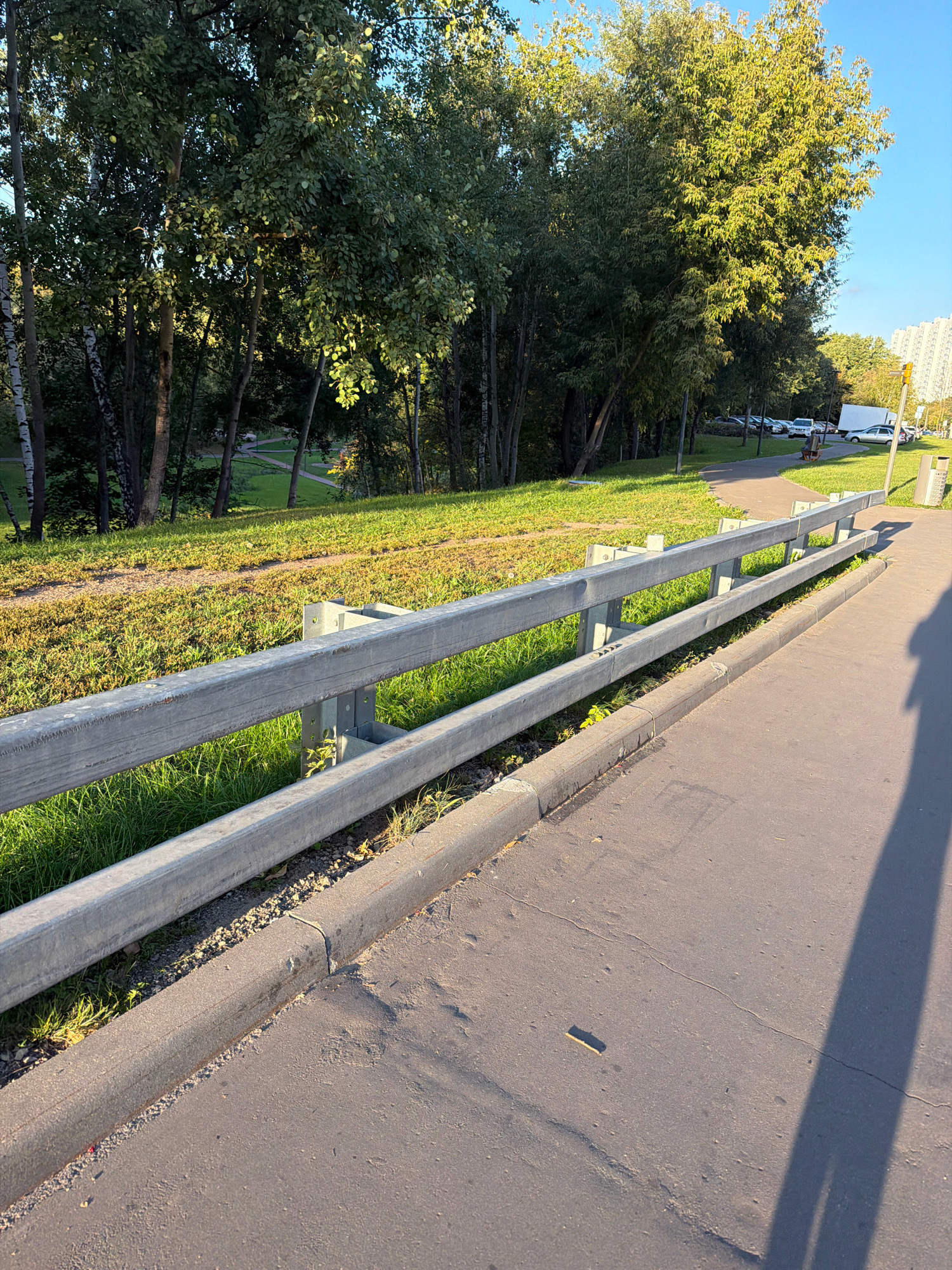 A low-angle, medium shot captures a paved walkway in Moscow, Russia, bordered by a silver metal guardrail and a grassy embankment. Sunlight illuminates the scene, casting long shadows from the guardrail and a nearby object onto the walkway. The guardrail, constructed of two horizontal metal bars supported by vertical metal posts, runs along the edge of the walkway. The walkway itself is asphalt with visible cracks and is bordered by a concrete curb. Beyond the guardrail, the grassy embankment slopes upward, dotted with patches of dry grass and small green plants. Further back, a dense stand of trees forms a natural backdrop, with sunlight filtering through their leaves. In the distance, a few parked cars, a white truck, and a tall, modern apartment building are visible. The sky above is clear and blue.
