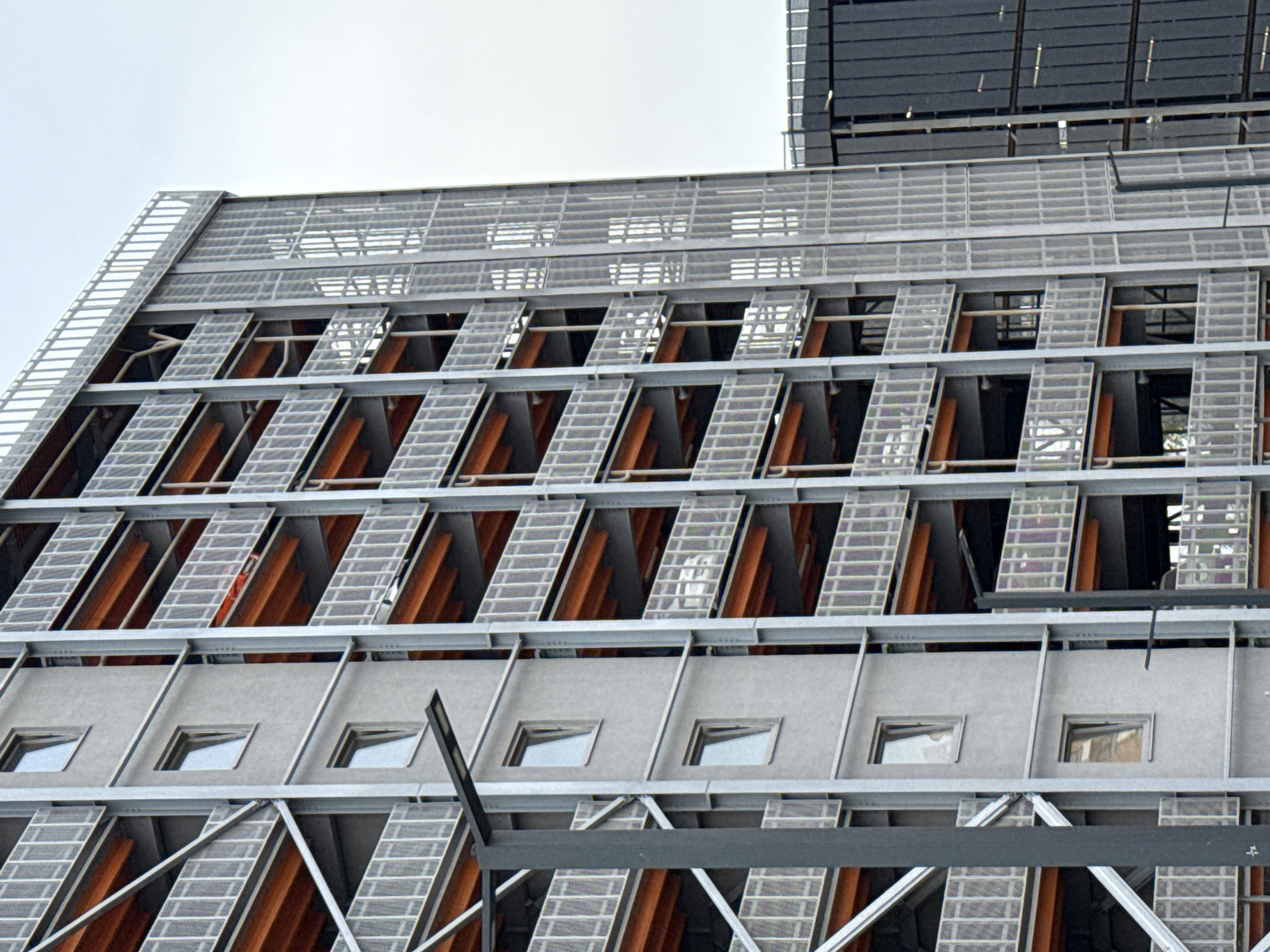 The image shows a multi-story building with a modern, utilitarian design, featuring concrete facades, exposed wooden beams, and metal railings on balconies or parking levels. The building has a series of uniformly spaced windows, some of which are slightly ajar. A street lamp is visible on the left side of the image, attached to a dark grey pole. To the right of the primary building, another structure with dark, vertical panels is partially visible. The sky is overcast and pale. There are no people or vehicles clearly discernible in the image. The overall impression is that of an urban architectural structure, possibly a residential building or a parking garage, with a functional and contemporary aesthetic. The specific location in Moscow, Russia, cannot be determined from the visual information provided.