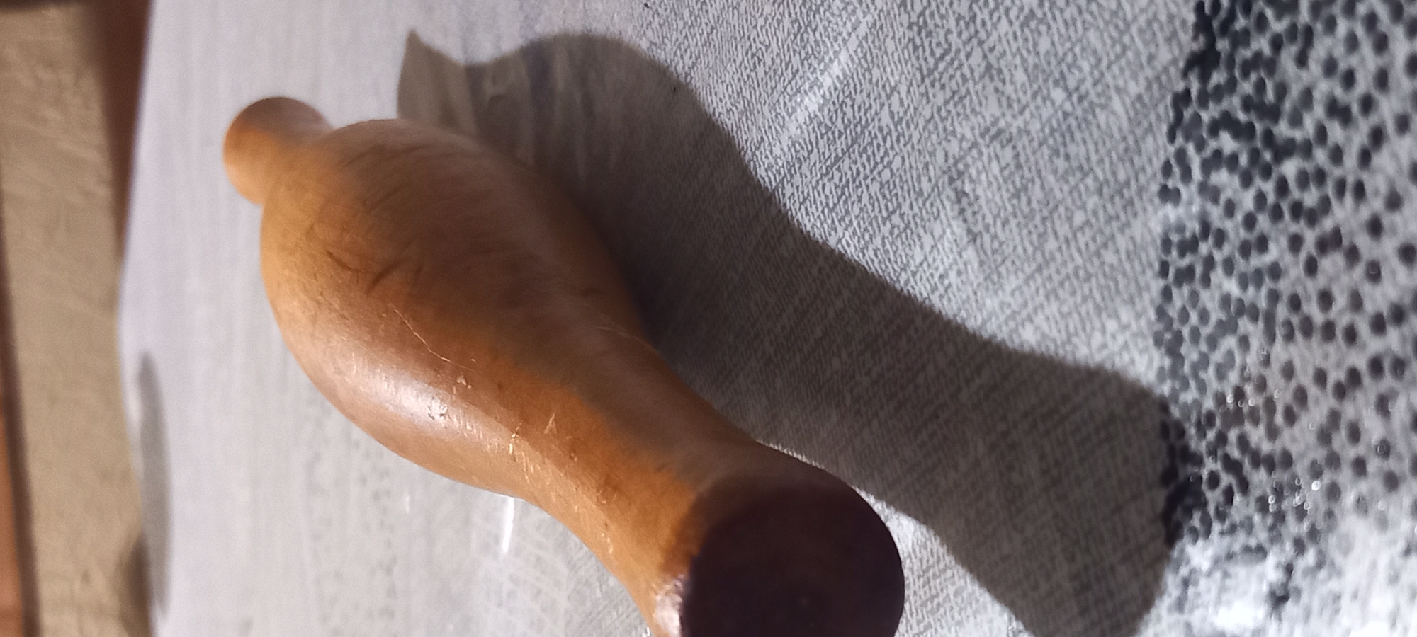 A light brown, elongated, carved wooden object, resembling a pestle or a handle, is positioned diagonally on a light gray patterned fabric surface. The object has a smooth, somewhat reflective finish with visible minor surface imperfections and scratches. Its broader, darker end is situated in the lower-left foreground, tapering towards a narrower, lighter end in the upper-right midground. A prominent shadow is cast by the object onto the fabric, extending towards the lower-left, indicating a light source from the upper right. The fabric exhibits a woven texture with a distinct, darker speckled pattern along its lower edge. The background is out-of-focus, showing blurred warm brown and beige tones, suggesting a wall or furniture. No individuals are present. The scene is static and devoid of active engagement, depicting a still life in Kryvyi Rih, Ukraine.