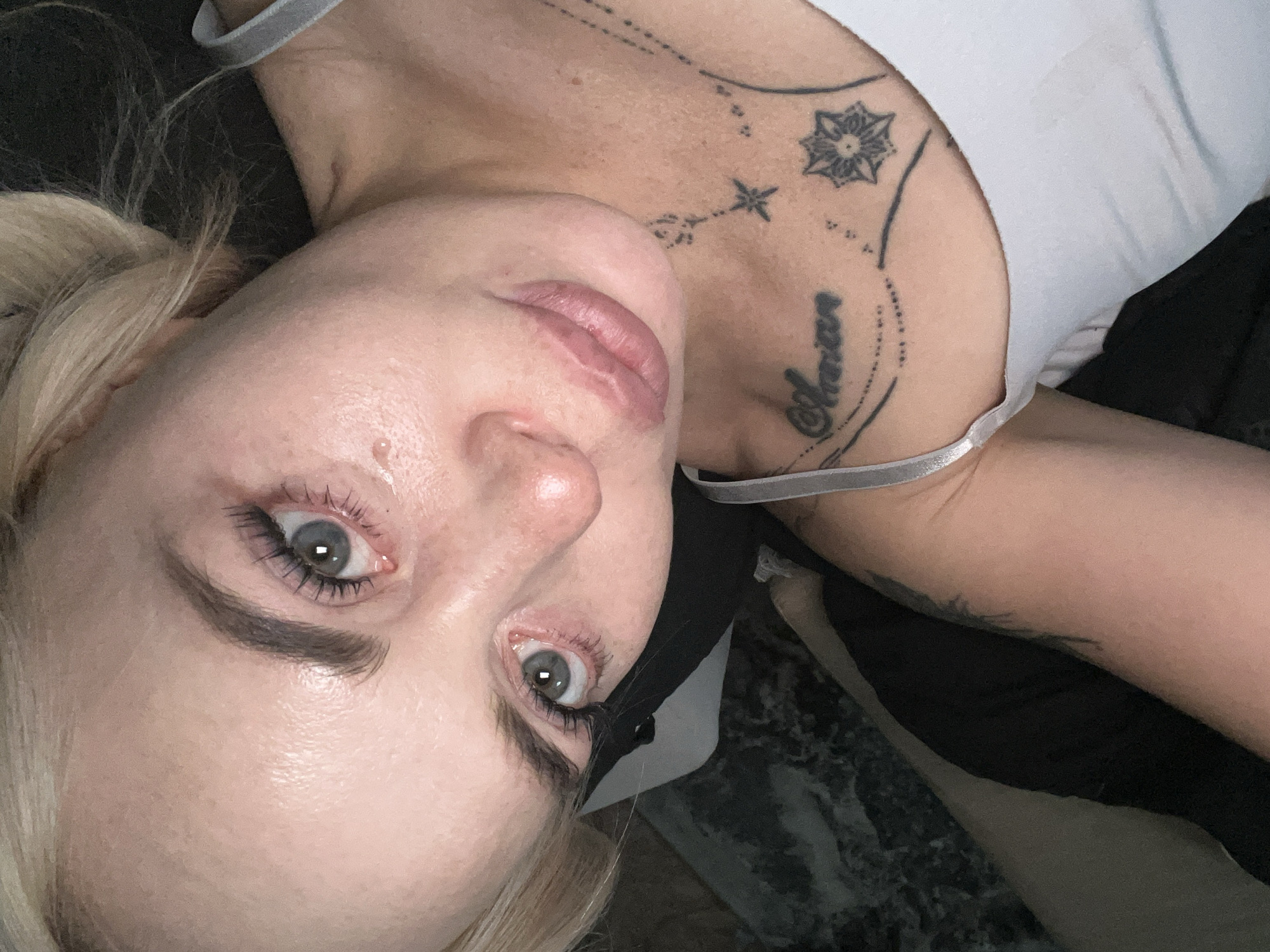 A close-up, overhead, rotated image shows a person's face. The person has fair skin, light blonde hair pulled into a ponytail on the left side of their head, and blue-gray eyes. They are wearing eye makeup, including mascara. The person's lips are colored with a matte lipstick.
Visible on the person's neck and upper chest are several tattoos. One tattoo is a word in cursive script, "Adrian," on the right side of the neck. There are also celestial-themed tattoos, including a starburst and a constellation-like pattern. The person is wearing a white tank top with a gray strap visible across their collarbone. A black mask is partially obscuring their chin and lower face.
The lighting suggests it is likely daytime, possibly indoors. The person's expression could be interpreted as pensive or resting. The background is dark and textured, possibly fabric.