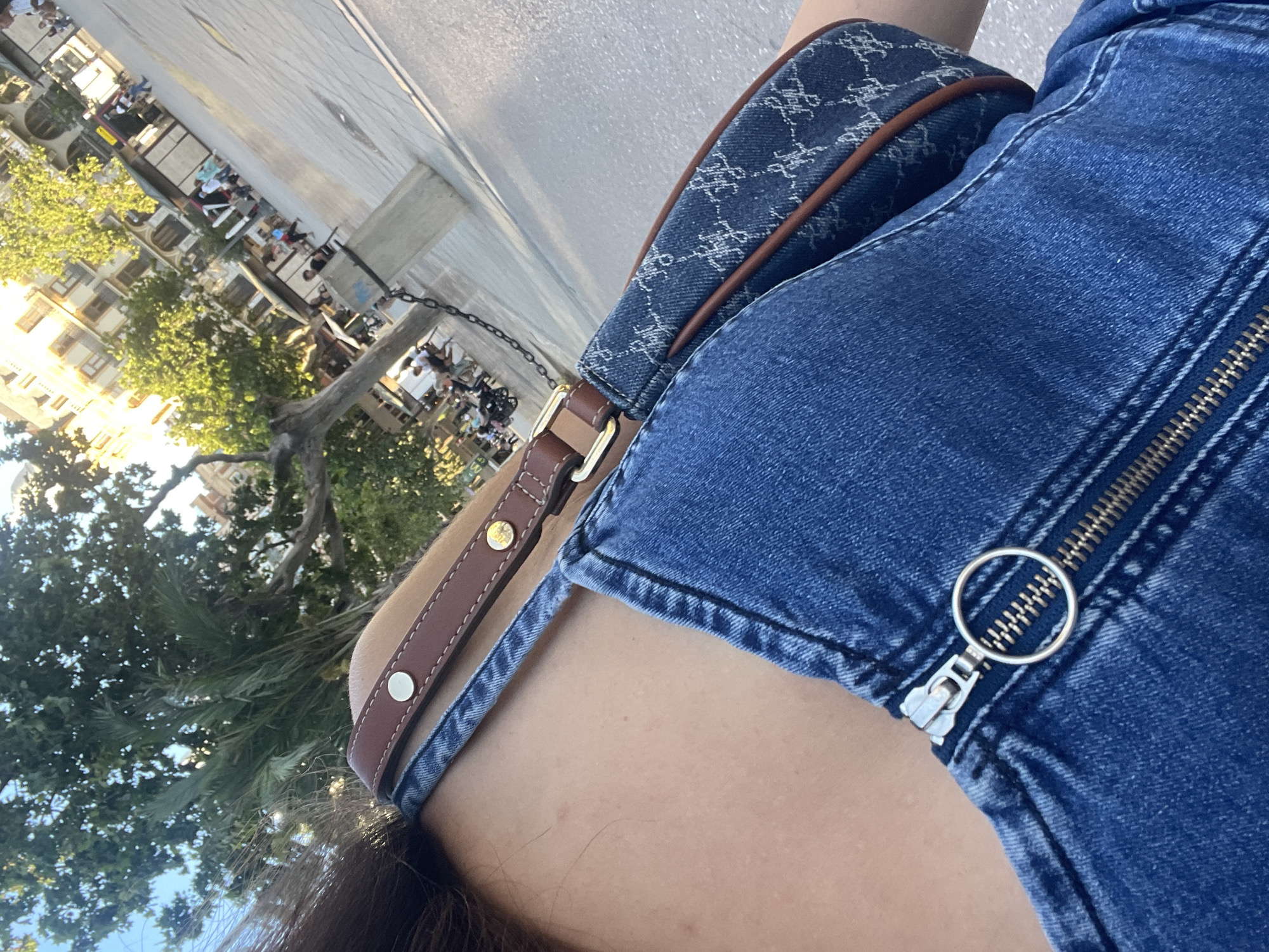 A high-angle, close-up shot depicts a person wearing a blue denim garment with a visible zipper and a handbag with a patterned blue fabric and brown leather strap. The strap is positioned across the person's shoulder. The background features out-of-focus trees and buildings, and appears to show a street scene in the city of València, Spain. The light suggests an outdoor setting during daytime. There are trees and some buildings are visible.