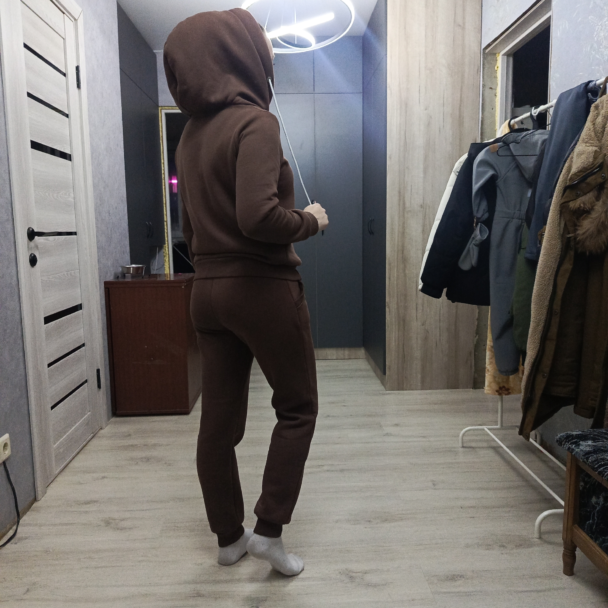 A person wearing a brown hooded tracksuit stands with their back to the camera in what appears to be a hallway or entryway in Markovskiy, Russia. They are wearing light-colored socks and casually holding the drawstring of their hoodie.
The indoor setting features light-colored laminate flooring and textured grey wallpaper on the left wall. A light wooden door with black horizontal inserts and a black handle is visible on the left. Further down the wall, a dark brown wooden cabinet or chest holds a silver bowl on its top surface. A white power outlet with a black cord is plugged in near the floor on the left.
On the right side of the image, a white metal clothing rack is laden with various garments, including a grey jacket, a black and white jacket, a dark jacket, and a brown faux fur or sherpa coat. Behind the clothing rack, an unfinished door frame with visible insulation foam suggests a recent renovation or construction. A wooden bench or ottoman with a dark patterned cushion is partially visible on the far right.
A modern, circular LED light fixture is installed on the ceiling. In the reflection of a dark cabinet or mirror in the background, a gold-framed mirror and a distant pink light can be discerned. The scene depicts a functional and contemporary residential interior. The person's expression is not visible, and there is no discernible text in the image.