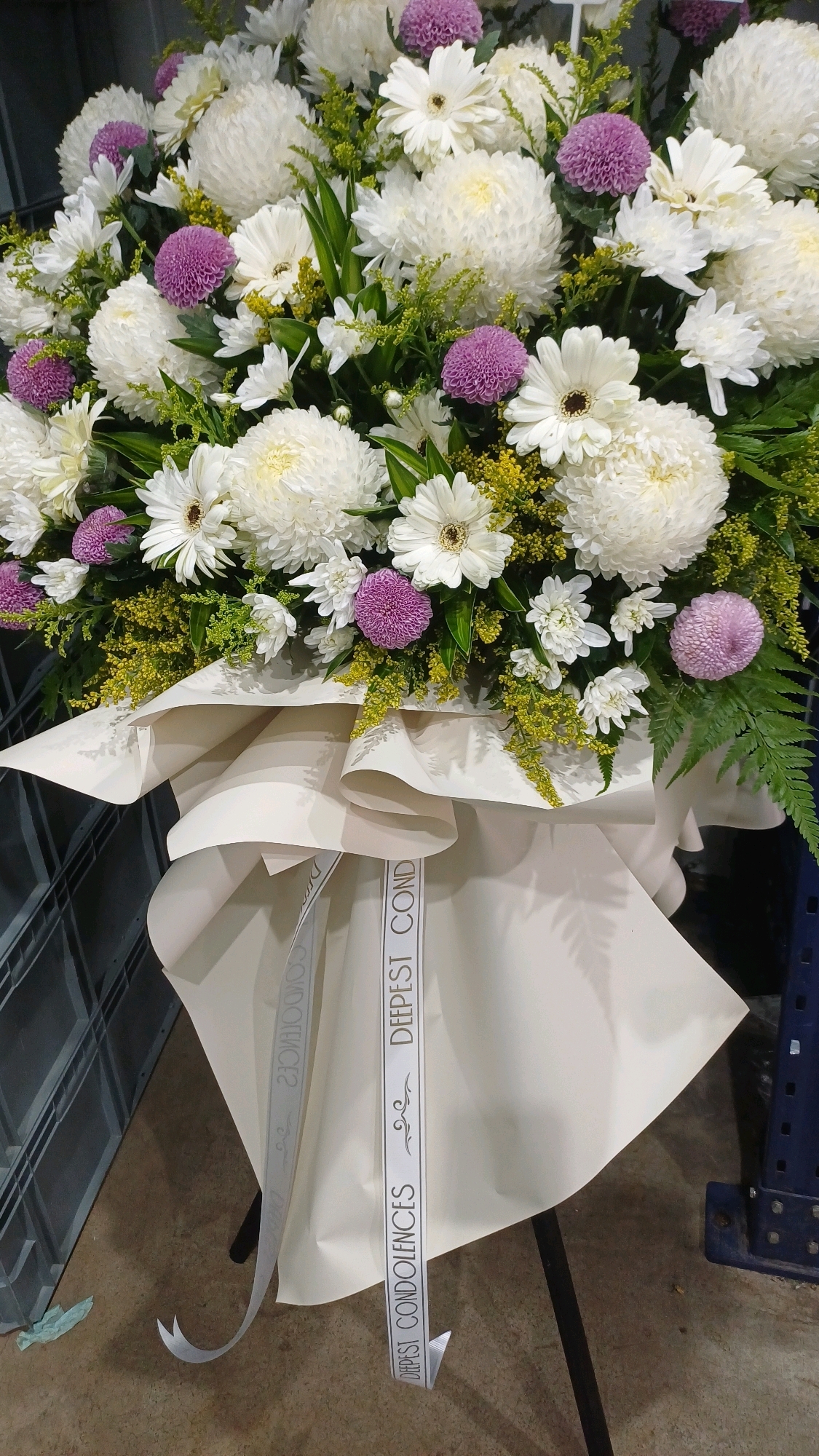 A close-up, eye-level shot shows a large floral arrangement of white and purple chrysanthemums and white daisies, with lush green foliage. The flowers are displayed in a cream-colored paper wrap, tied with a ribbon that reads "DEEPEST CONDOLENCES" in black lettering. The arrangement is situated in a warehouse-like environment, indicated by the presence of industrial storage racks and concrete flooring. The overall impression is of a formal bouquet, possibly for a funeral or memorial service. No people are visible in the image, and no specific location in Kuala Lumpur, Malaysia, can be identified.