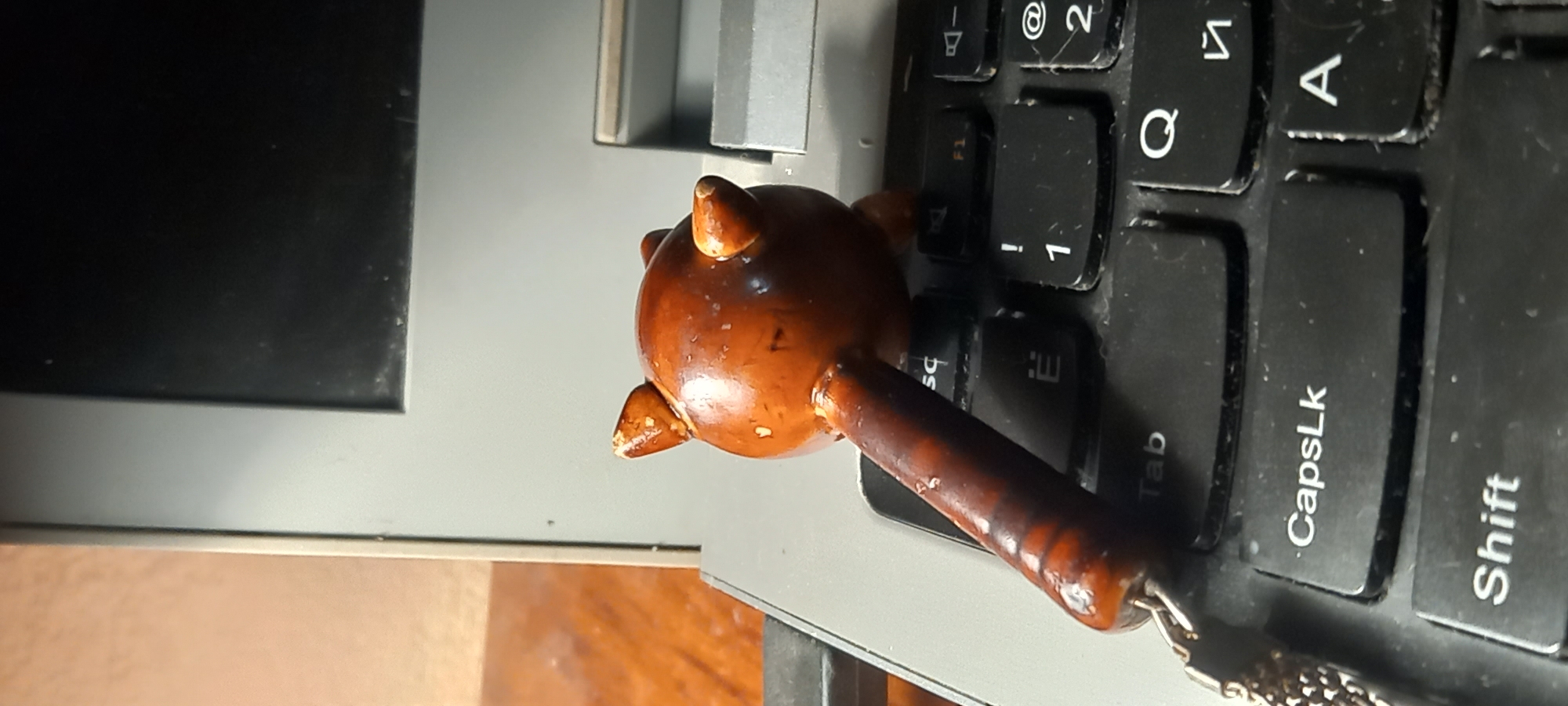 The media file displays a small, dark brown, wooden or resin object resembling a miniature mace or flail, positioned on a black laptop keyboard. The object features a rounded, spiked head and a ribbed handle, with a silver-colored chain attached to its base, suggesting it is a keychain or decorative charm. Its surface has a glossy finish and some minor wear marks.
The object rests over the laptop's "Tab," "CapsLk," and "Shift" keys, with part of its head obscuring the "Q" and "1" keys. The black keyboard features white Latin and Cyrillic alphanumeric characters. Visible keys include "Q," "A," "Tab," "CapsLk," "Shift," "F1," "1," "2," "!" and "@", along with other partially visible letters. There is noticeable dust and some grime accumulated on and between the keyboard keys, indicating the laptop's regular use.
The laptop screen bezel, black and seemingly made of plastic, is visible in the upper left, while the laptop's metal hinge mechanism is partially seen towards the top center. Below the laptop, a portion of a wooden or brown-colored surface, likely a desk or table, is visible. The overall setting appears to be an indoor workspace. The image is rotated 90 degrees clockwise from its natural orientation, showing the laptop on its side. The lighting is likely artificial, creating some reflections. The location context provided is Kryvyi Rih, Ukraine.