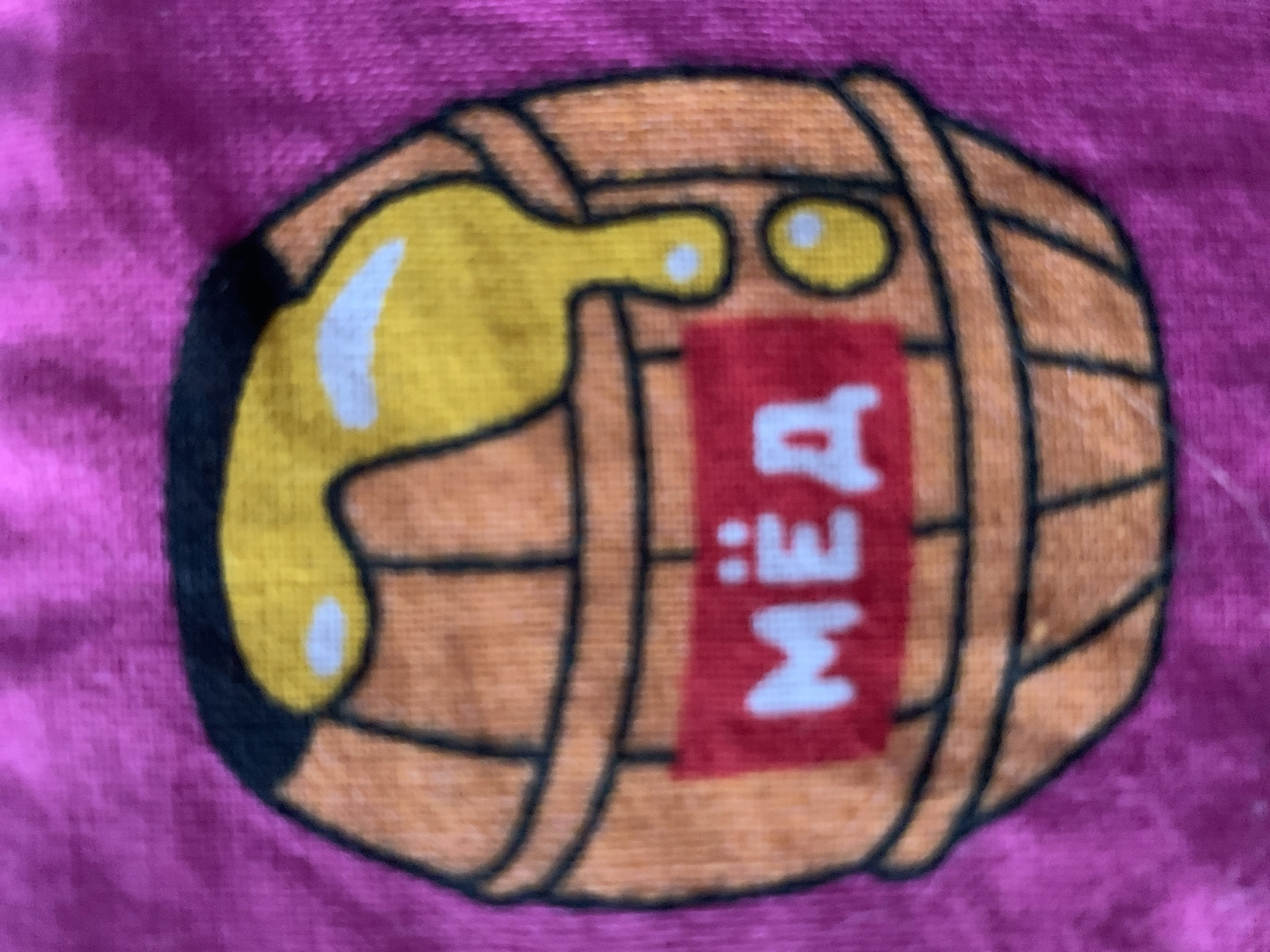 The image shows a close-up of a drawing on fabric. The drawing depicts a cartoon barrel with a yellow liquid, possibly honey or syrup, pouring out of it. The barrel is depicted with wooden staves and hoops. A red label with white text is attached to the barrel. The text on the label appears to be "MEIA", with the letters possibly reversed or in a stylized font. The background is a solid pink color, suggesting the fabric is a piece of clothing or textile. There are no people, animals, or specific environmental details visible. The focus is entirely on the illustrated barrel and its label. The lighting is even, and there are no strong shadows, indicating a neutral indoor setting. The image is slightly blurry, which affects the sharpness of the details.
