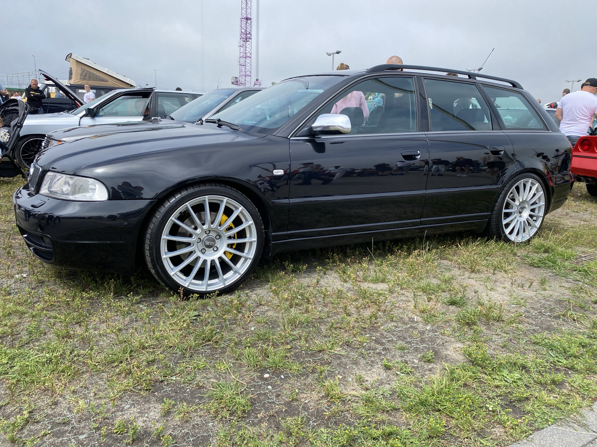 The image captures a black Audi station wagon parked on a gravel and grass lot under an overcast sky. The car features silver multi-spoke rims with yellow brake calipers and a roof rack. In the background, other cars are visible, including a silver SUV with a pop-top camper and a red sports car. Several people are present, some standing and observing the vehicles. The setting appears to be an outdoor car show or gathering. The presence of a purple tower structure and the style of the vehicles suggest a location like the Amber Expo exhibition center in Gdańsk, Poland, which frequently hosts automotive events.
