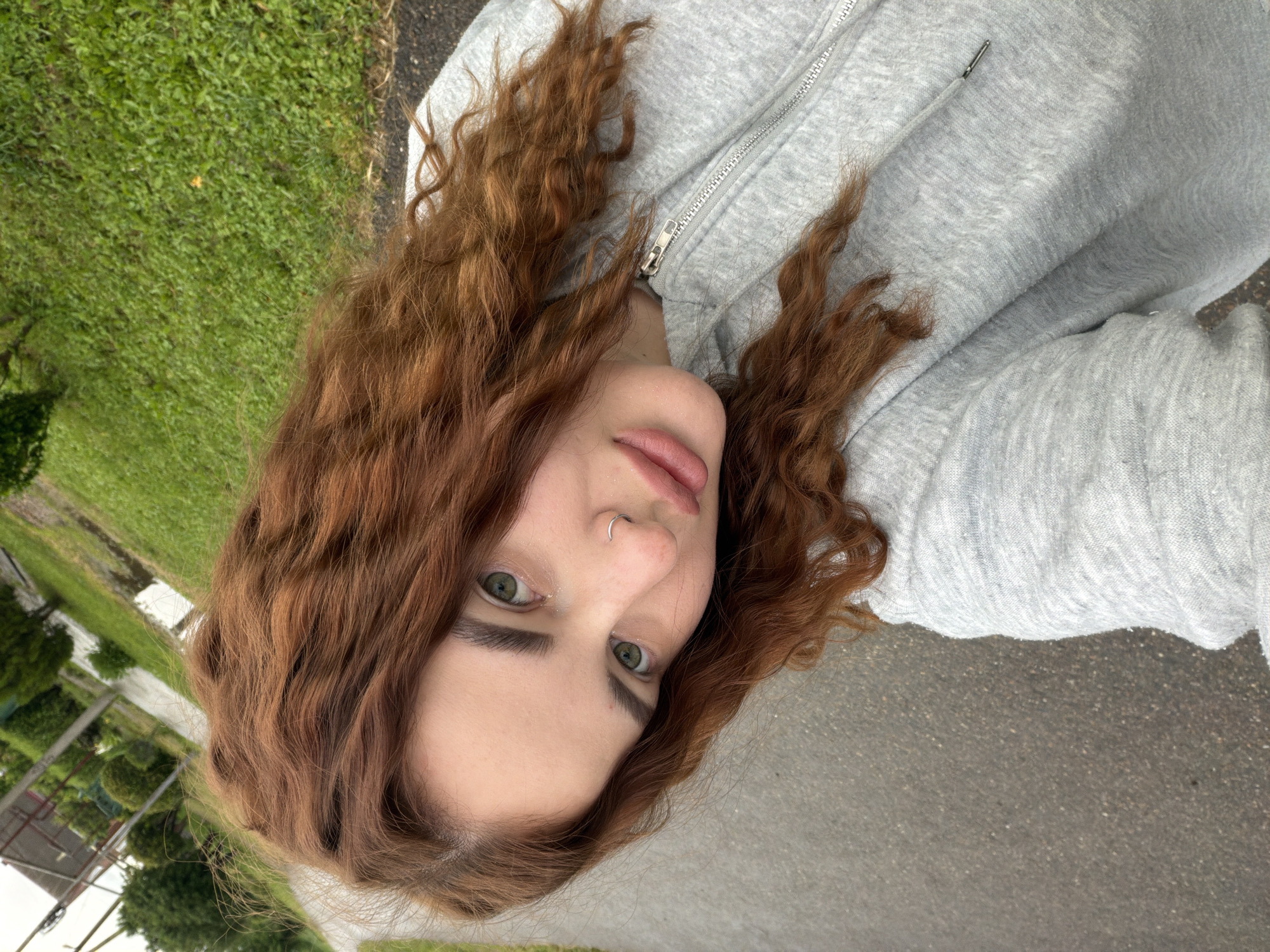 A woman with curly auburn hair, green eyes, and a nose ring is lying on gray asphalt wearing a gray zip-up hoodie. Green grass and trees are visible in the background. The location is likely near Novopokrovskaya, Russia.