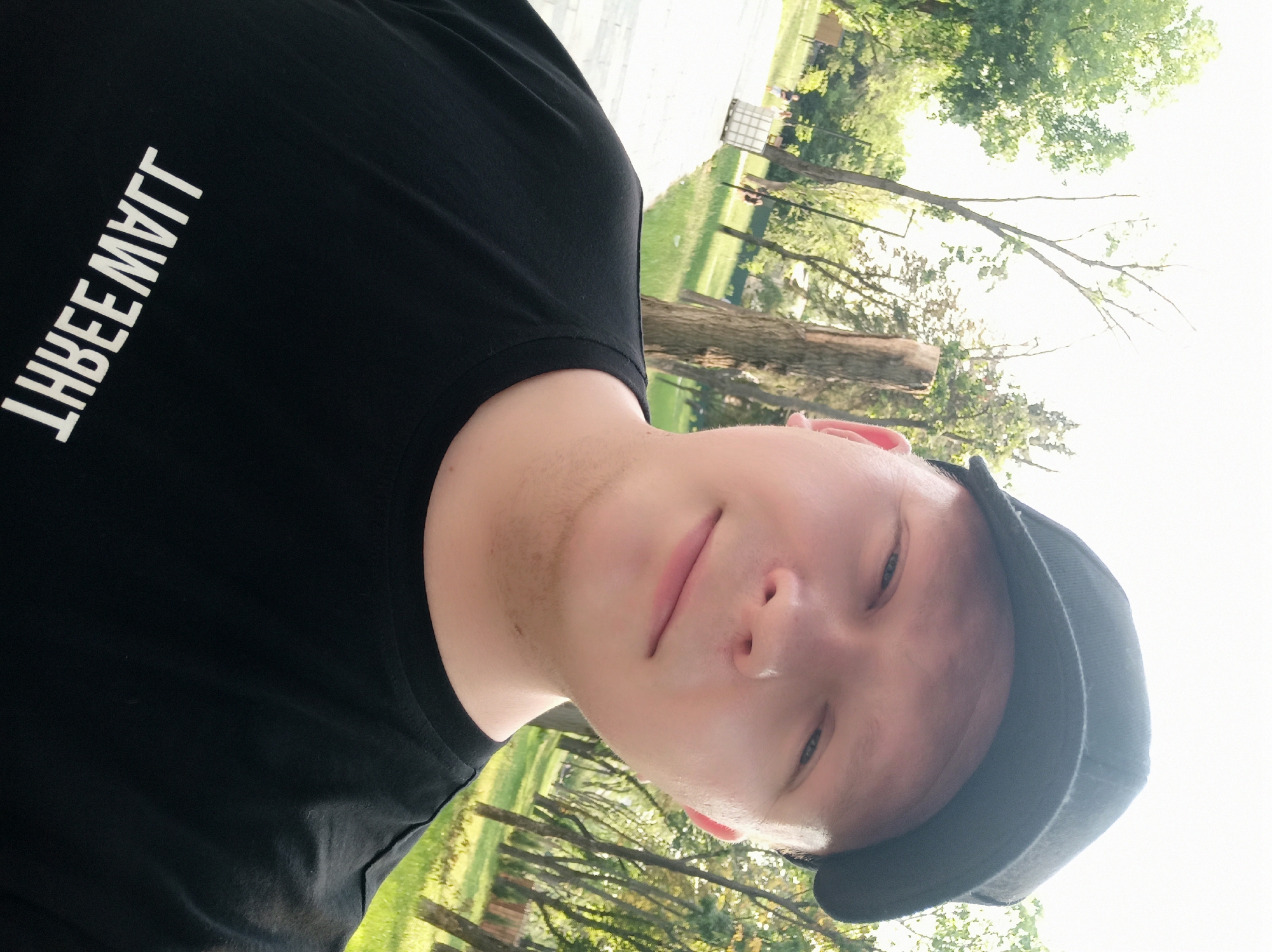 A young, fair-skinned individual wearing a black baseball cap and a black t-shirt with white lettering that reads "THREE WALL" backwards, is taking a selfie outdoors. The person is looking at the camera with a slight smile. In the background, a paved walkway runs through a grassy park with numerous trees. Some people can be seen walking in the distance, and there is a white industrial-style container on the left side of the walkway. The sky is bright and appears to be sunny.