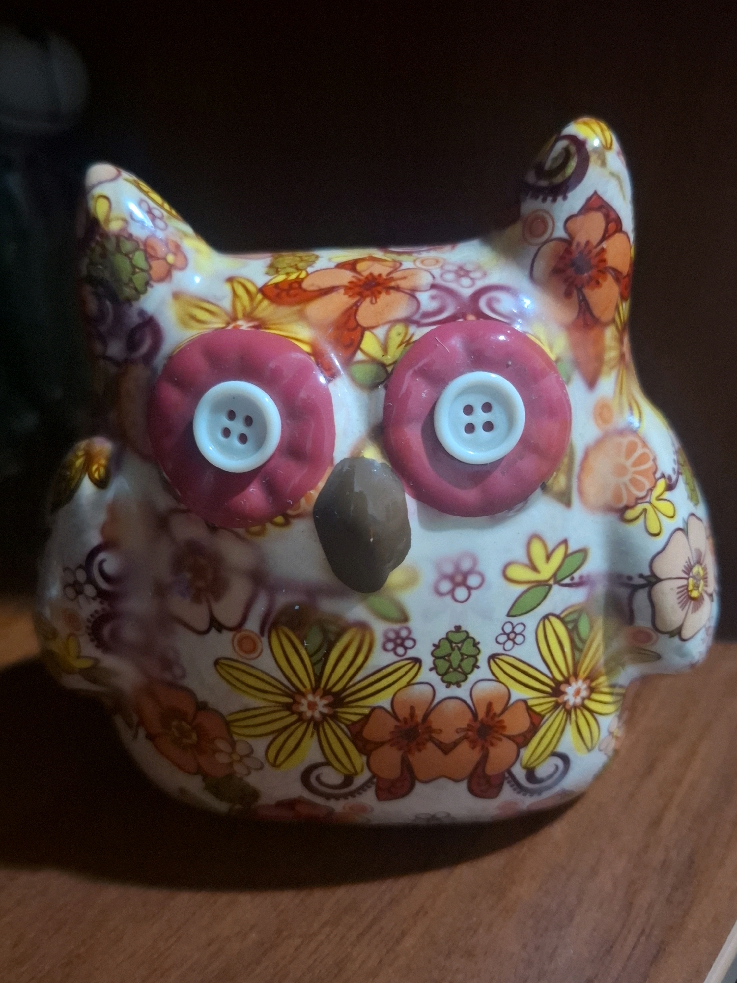 A close-up, eye-level shot shows a decorative ceramic owl figure. The owl is predominantly white with a colorful floral pattern featuring orange, yellow, pink, and green flowers and swirls. It has two large pink button eyes, each with a white four-hole button centered on it. A small, brown, oval-shaped object serves as its beak. The owl is positioned on a wooden surface, likely a table or shelf, with a dark, blurred background. The lighting suggests an indoor setting. There is no discernible connection to the city of Almaty, Kazakhstan.