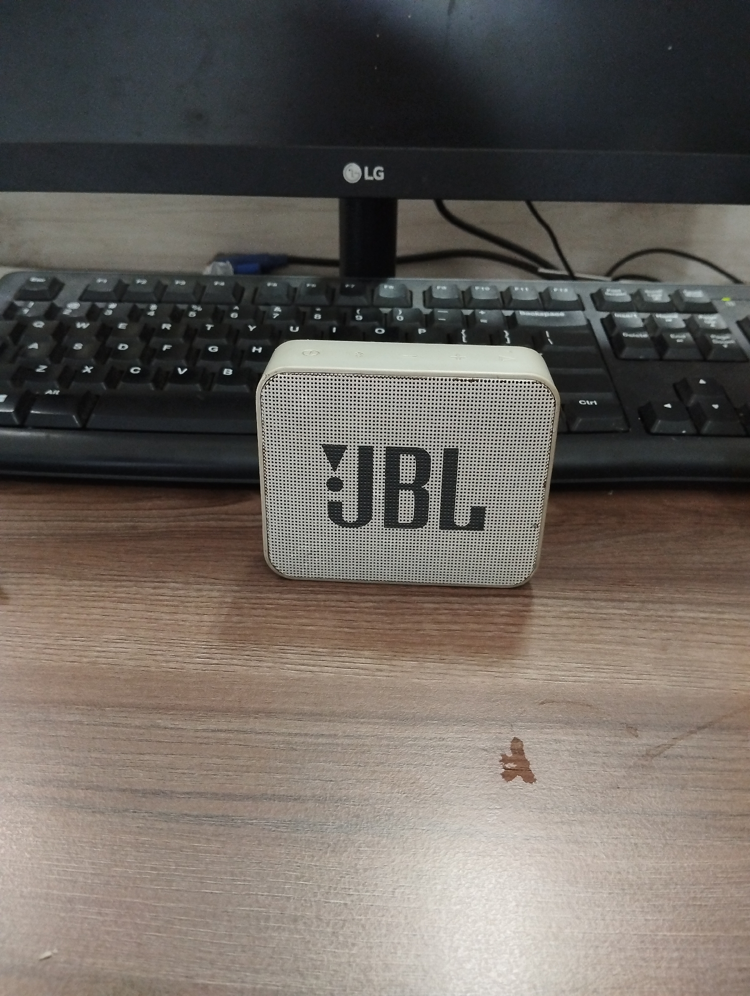 An eye-level shot shows a white JBL portable speaker placed in the foreground on a wooden desk. Behind the speaker, a black computer keyboard is visible, with the monitor of a black LG computer screen in the background. The desk surface has a visible wood grain pattern. The overall scene appears to be a home or office workspace. The location is described as Kelapa Dua, Indonesia, although there are no specific visual cues in the image to confirm this.