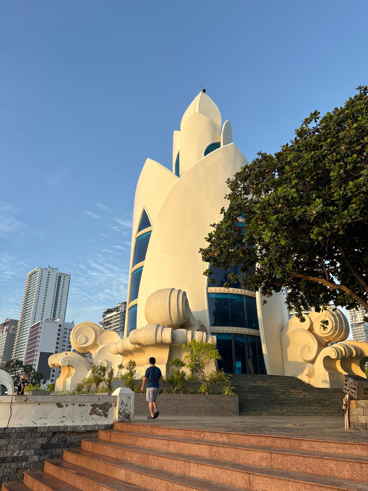 This is a photograph taken in Nha Trang, Vietnam, featuring the iconic Dragon Beach Gate structure. The image is captured during daylight hours under a clear blue sky. In the foreground, a man in a blue t-shirt and grey shorts walks up a set of reddish-brown tiled stairs. To his right, a large, leafy green tree partially obscures a portion of the white, curved building. The building itself has a unique, organic design, with prominent blue-tinted windows. At the base of the building are stylized, wave-like sculptures and low-lying green plants. In the background, several tall, modern buildings rise into the sky, indicating an urban setting. The overall scene suggests a warm, sunny day at a coastal location in the city.
