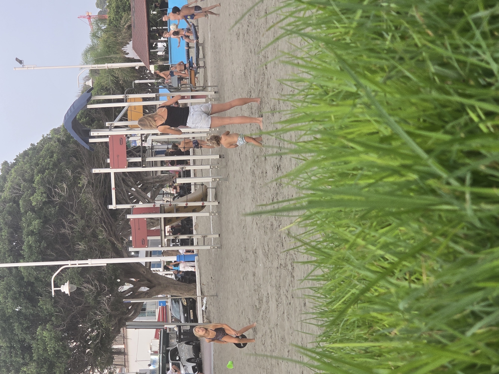This image depicts a public beach area with a playground in the background. In the foreground, blades of green grass partially obscure the scene. On the sandy beach, a young girl in a patterned swimsuit is actively playing with a paddle and ball. Further back, a woman in a black tank top and denim shorts stands with her back to the camera, watching a toddler in a diaper. Several other children and adults are scattered throughout the playground area, some appearing to be engaged in play or conversation. The playground itself features red and yellow structures, a blue slide, and metal supports. In the distance, a street with parked cars and buildings, including what appears to be a restaurant with outdoor seating, is visible. Palm trees and other lush vegetation are also present, typical of a coastal city like Málaga, Spain. The overall atmosphere suggests a casual, recreational setting.