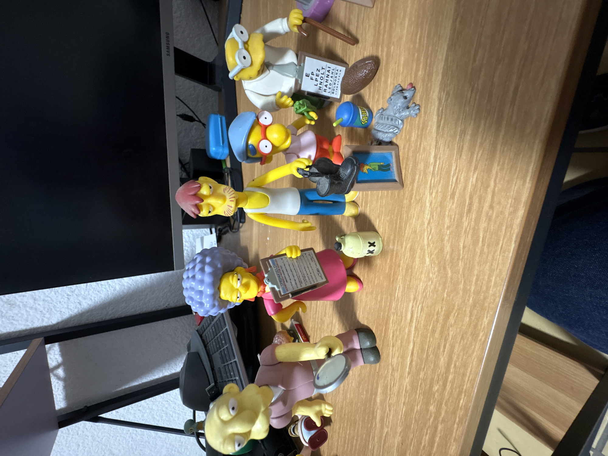 A collection of "The Simpsons" figurines and related accessories are scattered on a wooden table.
The figurines include:
* Dr. Hibbert holding an eye chart and a pointer
* Comic Book Guy holding a comic
* Sideshow Bob with pink hair, wearing blue jeans
* Patty Bouvier holding a clipboard
* Hans Moleman holding a magnifying glass
Accessories include:
* A gray rat
* A wooden frame depicting a character lying down
* A bottle labeled "SOULS"
* A gas tank labeled "XX"
* A can of Duff beer
* A turtle
* A blue box
In the background, there's a Samsung monitor, a shelf, and what appears to be a section of a blue chair. The setting is indoors.