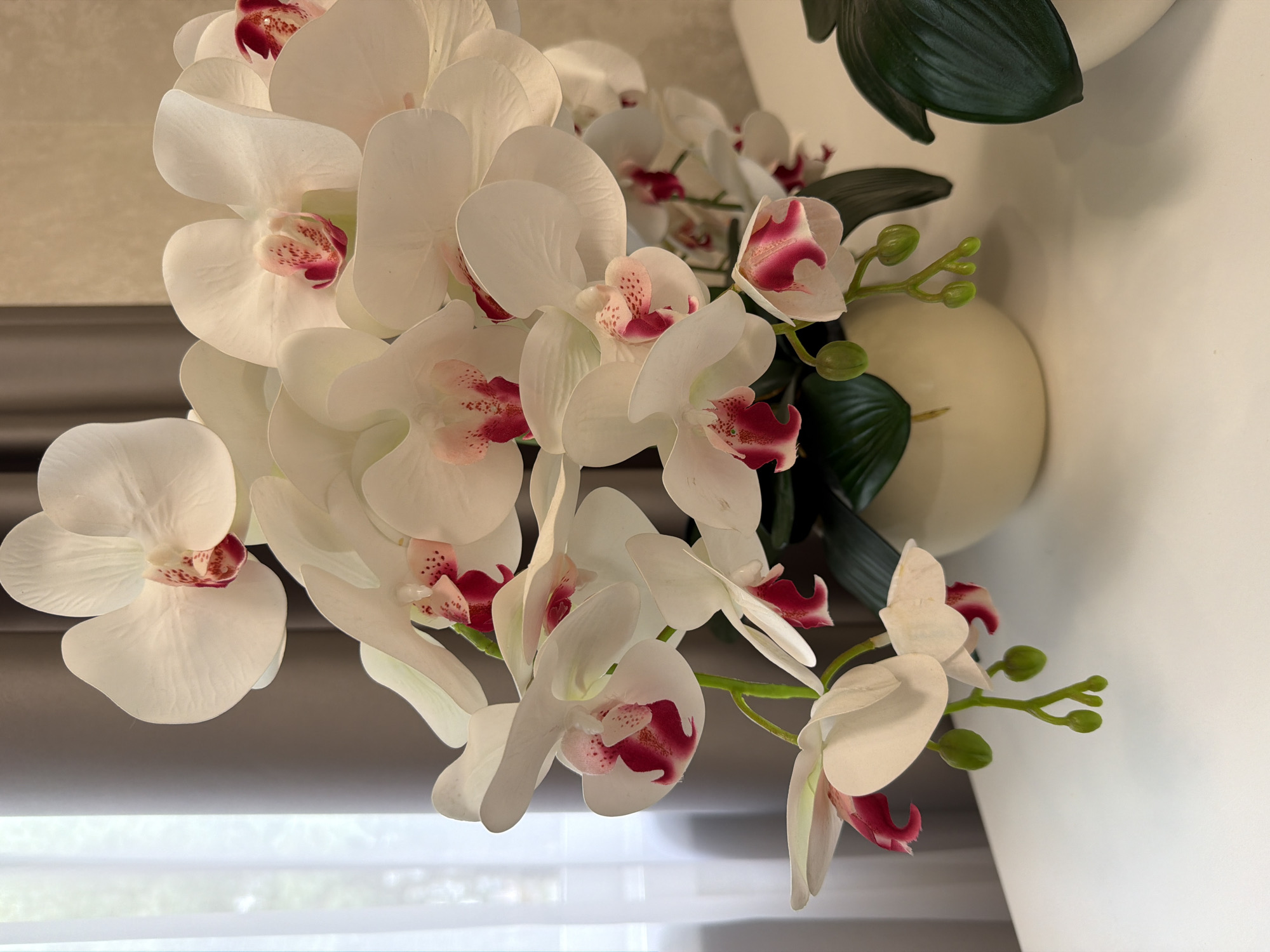 A close-up, eye-level, medium shot captures a bouquet of artificial white orchids with deep pink centers, arranged in a cream-colored ceramic pot. The orchids are densely packed, with some blooms facing forward, others to the side, and a few buds hanging down. The background is softly blurred, featuring a sheer white curtain to the left and a pale beige wall behind the arrangement. The orchids rest on a white, reflective surface, likely a table, with another out-of-focus, dark green plant visible on the right. The lighting is soft and diffused.