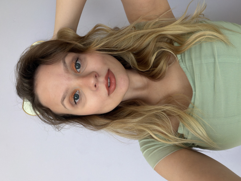 An adult female is depicted in a close-up, chest-up indoor portrait. She has light skin, blue eyes, and long, wavy hair with blonde highlights. Her hair is parted slightly to her left and flows over her shoulders. She wears orange eyeshadow and a reddish-brown lipstick. Two light-green, ear-shaped objects, possibly part of a headband or accessory, are partially visible behind her head. She is wearing a light green, short-sleeved top with a v-neck and textured fabric. Her right arm is raised, with her hand positioned near the back of her head. Her left shoulder and arm are partially visible. She looks directly into the camera with a neutral expression. The background is a plain, light grey wall. The scene is consistent with an indoor setting within a residence in Tel-Aviv, Israel; however, no specific location markers within the city are visible in the image.