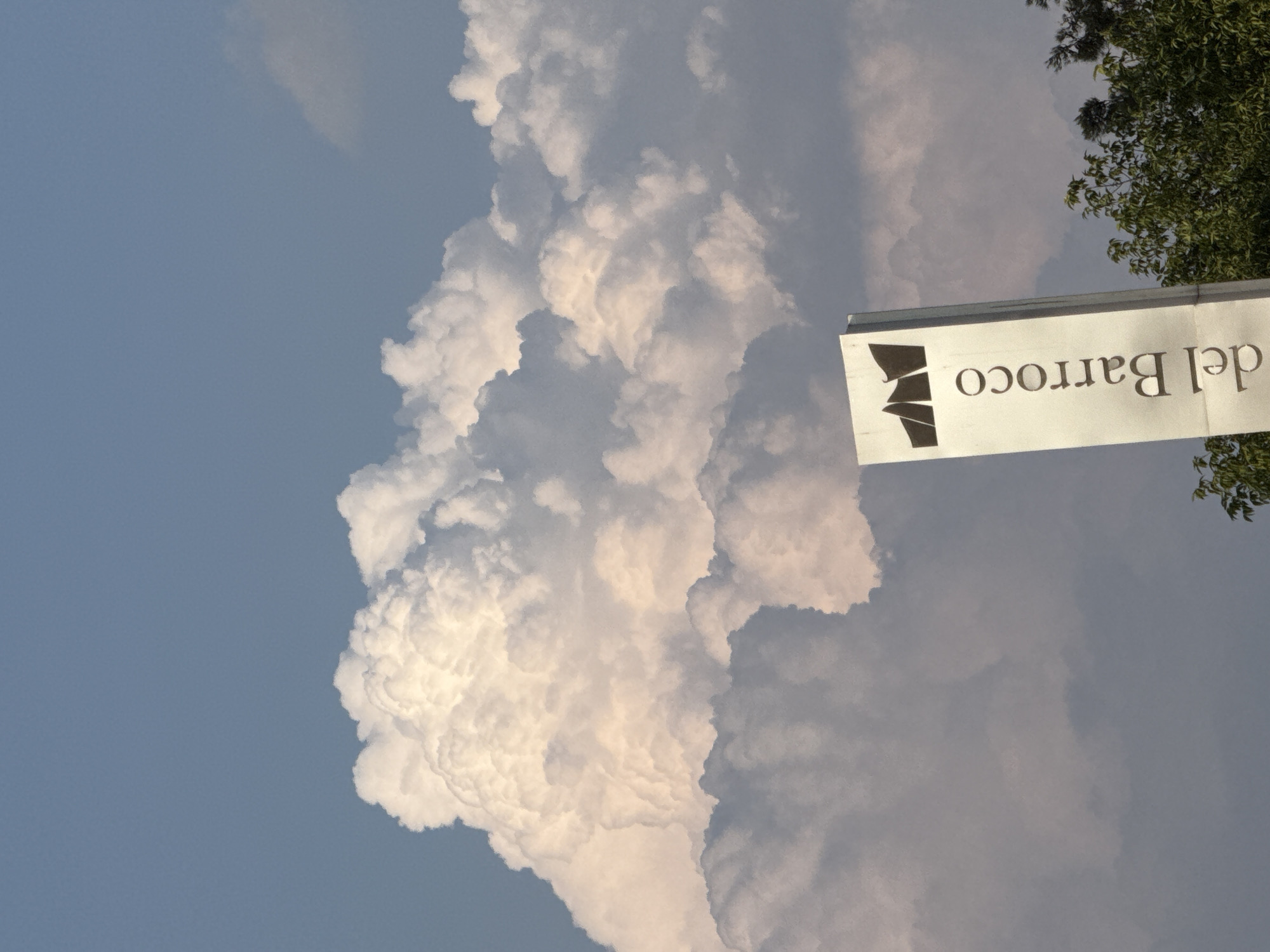 The image shows a sky scene with a large cumulus cloud formation, partially obscuring the blue sky. A sign, oriented horizontally, reads "del Barroco" and features a stylized logo resembling sails. Green foliage from a tree is visible near the sign. The location is likely San Bernardino Tlaxcalancingo, Mexico, given the sign's language (Spanish).