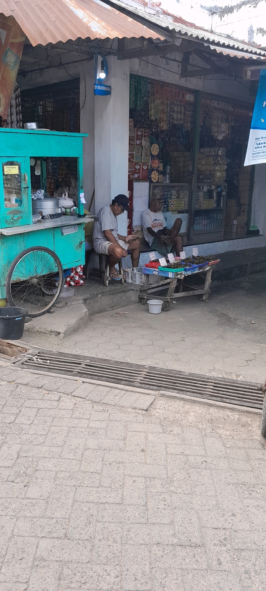 In Banyuwangi, Indonesia, two men are seen sitting outside a small store. One man, wearing a light grey t-shirt and shorts, sits on a stool and looks at his phone. The other man, in a white t-shirt with a graphic design, sits on the ground. In front of them is a wooden table with various items displayed, including what appear to be small packages or food items. A green stall is visible to the left, with a bicycle wheel leaning against it. The ground is paved with interlocking bricks, and a metal grate is present in the foreground. The store behind the men is stocked with numerous products, and a blue sign with white lettering that reads "O SEDERHANA" is attached to the building's exterior. The building has a rusty corrugated metal roof.