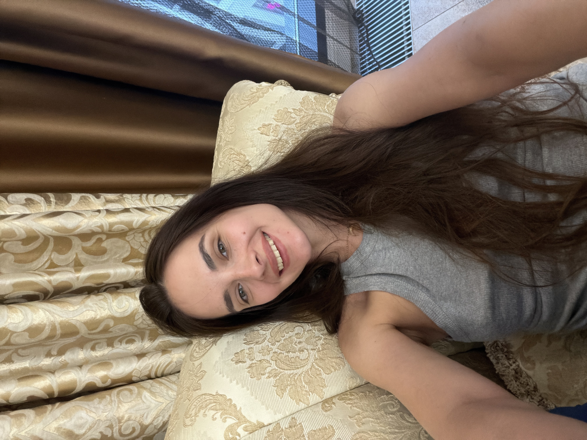 A young woman with long, dark brown hair is sitting on an ornate couch, looking at the camera and smiling. She is wearing a grey tank top and a delicate necklace. The couch has a cream-colored fabric with a repeating damask pattern in a lighter shade. Behind her, golden-brown curtains with a similar damask pattern are visible, along with a darker brown curtain to the right. The background also shows a glimpse of a window with a patterned screen. The lighting is soft, suggesting an indoor setting, possibly a living room. There is no indication of her location within a city.