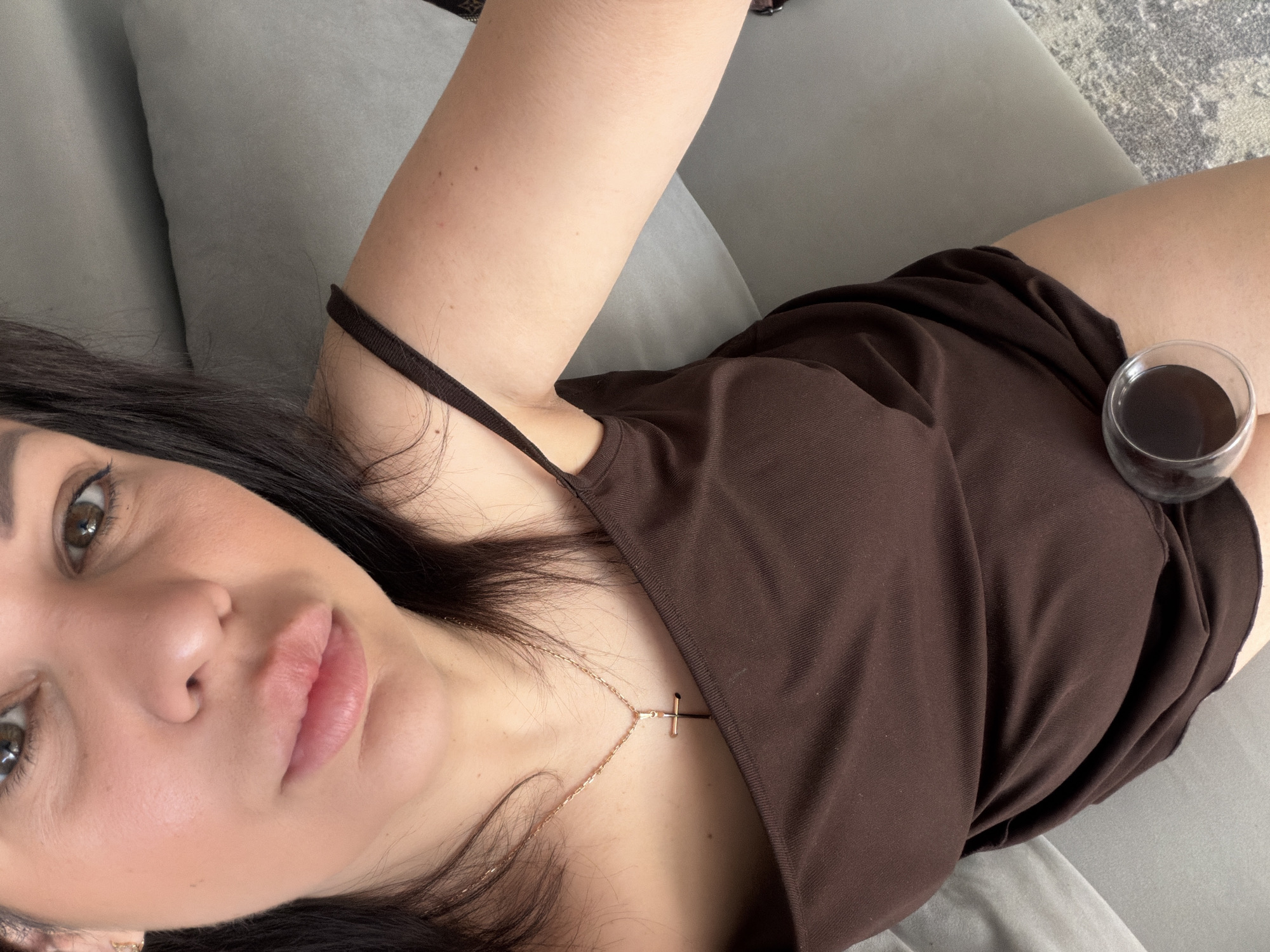 The image captures a close-up, overhead view of a female subject indoors in Sochi, Russia. The individual has dark hair and is looking directly into the camera. She wears a dark brown spaghetti-strap top, partially revealing her upper chest and left arm. A golden chain necklace with a cross pendant is centered on her chest. Her face exhibits minimal makeup, including eyeliner, and her lips are full and slightly parted. A clear glass containing a dark red liquid, consistent with wine, is positioned on or near her upper left thigh. The background features light grey upholstered furniture, likely a sofa or bed, and a textured grey patterned rug is visible on the floor in the lower right portion of the frame.