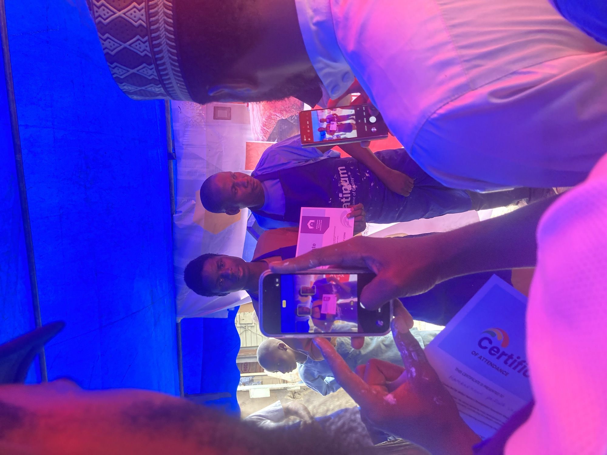 A photo captures a moment in Jos, Nigeria, where two individuals are receiving certificates, presumably for attendance. One man is wearing a "Platinum" t-shirt, while another dons a patterned hat. They are holding their certificates. Another person is photographing them with their smartphone. Someone else is also holding up a certificate to be photographed, suggesting multiple people are involved in this process. The setting is under a blue canopy, likely an outdoor event.