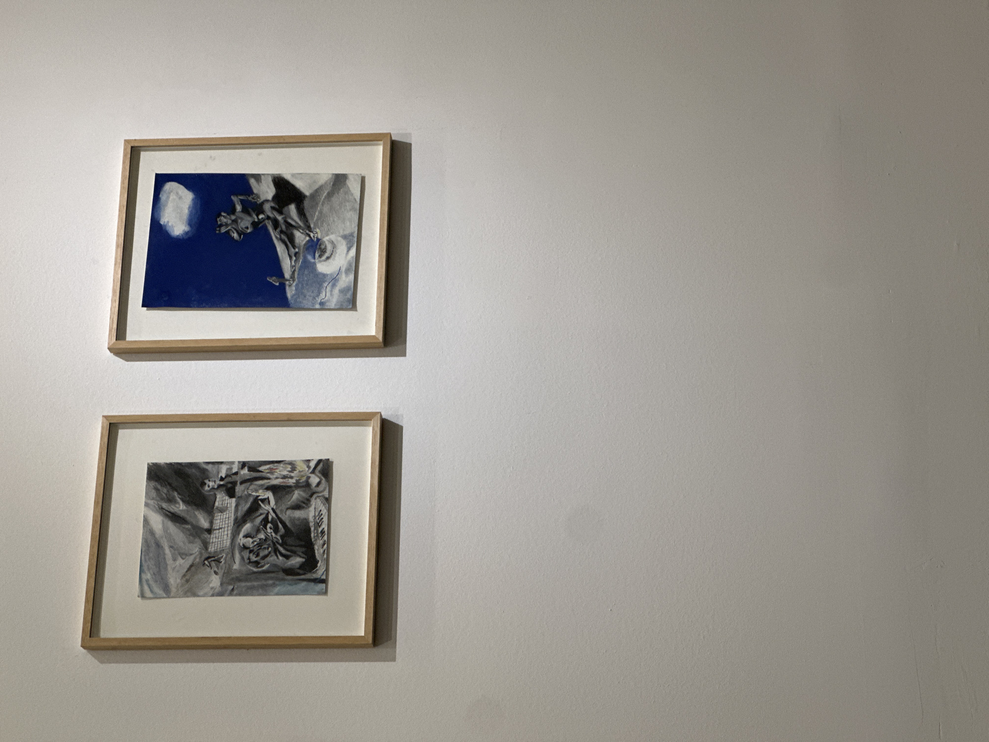 Two framed artworks hang on a light gray wall. The top artwork features a dark blue background with a lighter abstract design. The bottom artwork has a lighter gray background with a similar abstract design. The frames are a light wood color. The location is Tbilisi, Georgia.