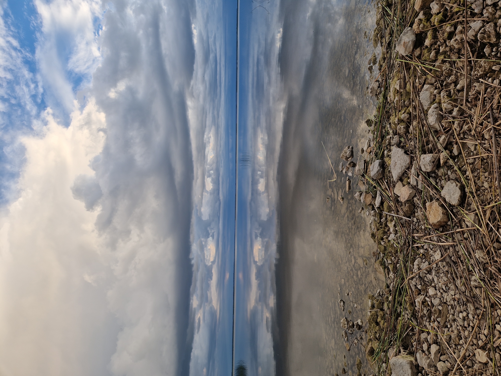 The image depicts a serene natural landscape with a calm body of water, likely a lake or the sea, under a dramatic, cloudy sky. The foreground is dominated by a stony shoreline covered with dry reeds and grass. The water's surface acts as a mirror, reflecting the textured clouds and the distant horizon, which appears as a thin blue line. Some sparse reeds or marsh grass are visible sticking out of the water near the left side. The overall impression is one of tranquility and a slightly overcast, possibly pre-storm atmosphere. The location is specified as the city of Īdeņa, Latvia.