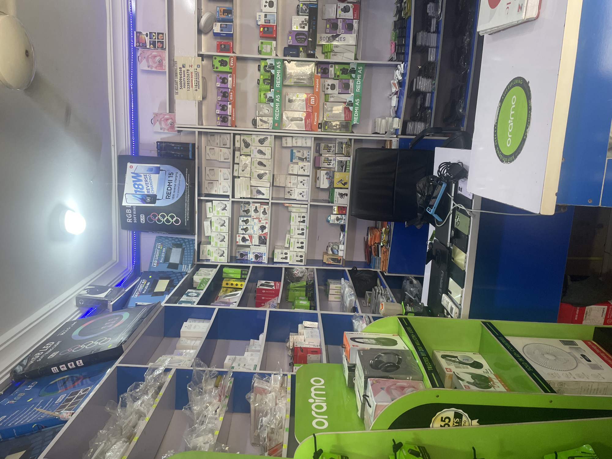 This media file depicts the interior of a mobile phone and accessories shop in Jalingo, Nigeria. The scene is filled with shelves packed with various electronic items, predominantly mobile phone accessories like chargers, earphones, and power banks. Prominently displayed are boxes for "Redmi A5" and "ORaimo" branded products, indicating these are key items in the store.
Several shelves are organized with smaller, individually packaged items, many of which appear to be chargers and cables. In the foreground, larger packages for "LED Light Kit" and "RGB Soft Ring" are visible. The store is well-lit, with a combination of overhead lighting and accent lighting on the shelves.
On the right side of the image, a portion of a blue and white counter is visible, with a large green display stand featuring the "ORaimo" logo. Text on the green stand highlights "Trusted by 200 Million+ Users" and "Sold in 60+ Countries." A ledger or notebook with "VIC DEELIGHT COMMUNICATION" and bank details is also visible, suggesting business operations are underway.
The overall impression is of a busy retail environment specializing in mobile electronics. The lighting and the abundance of products suggest it is daytime, and the clean, organized shelves imply the shop is operational and catering to customers. There are no people visible in the frame, focusing the viewer's attention solely on the merchandise and the store's layout.