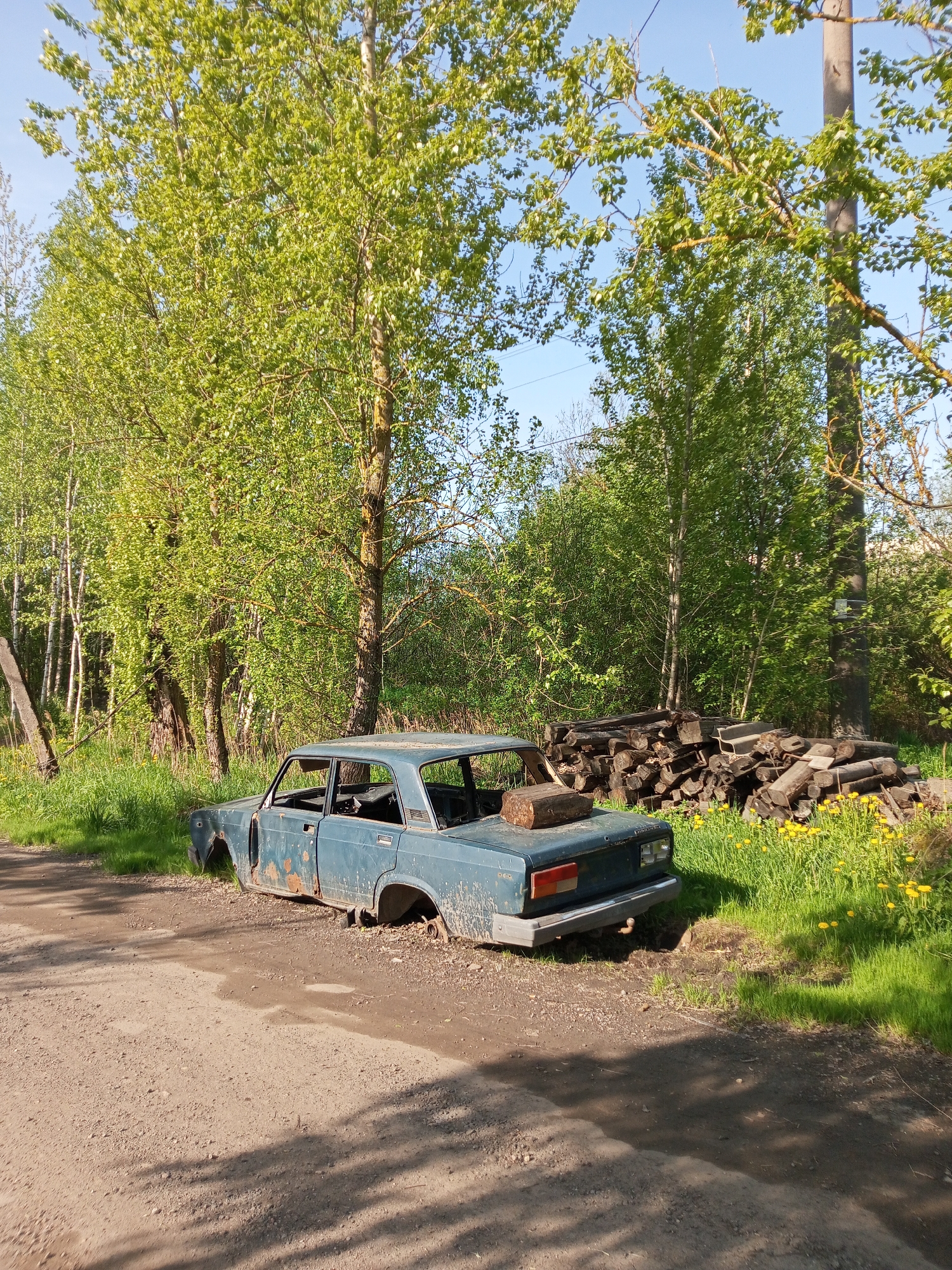 The image shows a derelict, blue, Lada car abandoned on the side of a dirt road. The car's doors are missing and the interior is stripped out. The car is rusting and partially submerged in the dirt. A log is resting on the trunk of the car. Adjacent to the car is a pile of chopped firewood. The scene is set within a rural environment, with trees bordering the road and patches of green grass and yellow dandelions. The sky is visible in the background and is a clear blue. Although the location is stated to be Kommunar, Russia, there are no specific details in the image to confirm the location. The scene appears to be in a state of neglect and abandonment.