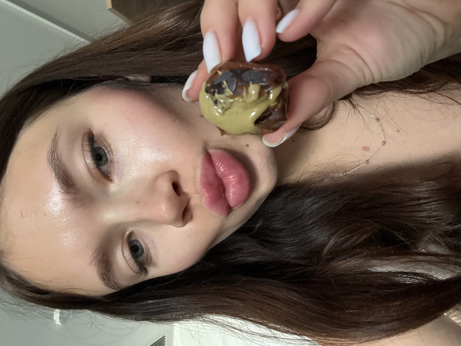 A woman with long brown hair and fair skin presents a chocolate-covered treat filled with a green substance. She holds it up near her lips with a manicured hand. Her eyes are blue-grey, and she wears light makeup. A delicate necklace adorns her neck. The image is taken from a low angle, giving a close-up view of her face and the confectionery.