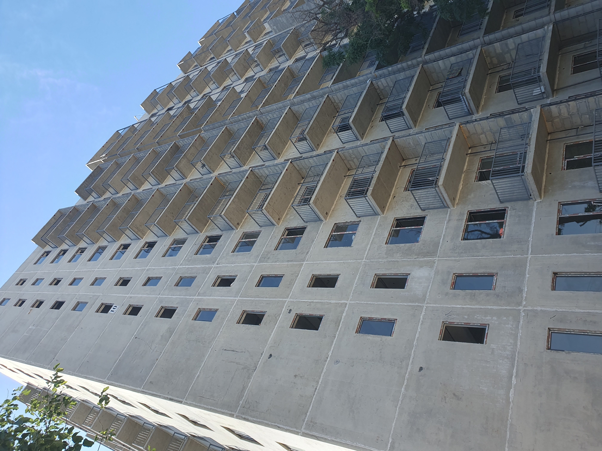 This is a low-angle shot of a multi-story apartment building under construction in Tashkent, Uzbekistan. The building is made of concrete and appears unfinished, with many windows unglazed and balconies bare. The sky is clear and blue, with some tree branches visible on the left side of the frame. The building's facade is a uniform gray concrete, with horizontal lines indicating floor divisions. There are metal railings on each balcony. The overall impression is one of ongoing construction and development in the city.