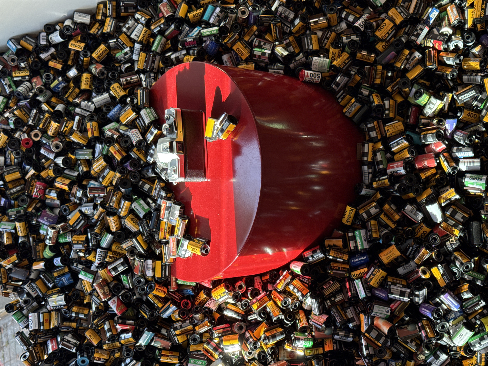 A high-angle shot depicts a dense collection of 35mm film cartridges, mostly Kodak brands, filling a container. The dominant color scheme is yellow and black from the film cartridges. A red object with a curved shape occupies the center of the frame. On top of it, a small, silver camera is placed. The scene is bathed in bright light, creating highlights and shadows throughout the collection of film cartridges. There is no mention of Belgrade, Serbia, or other location details.
