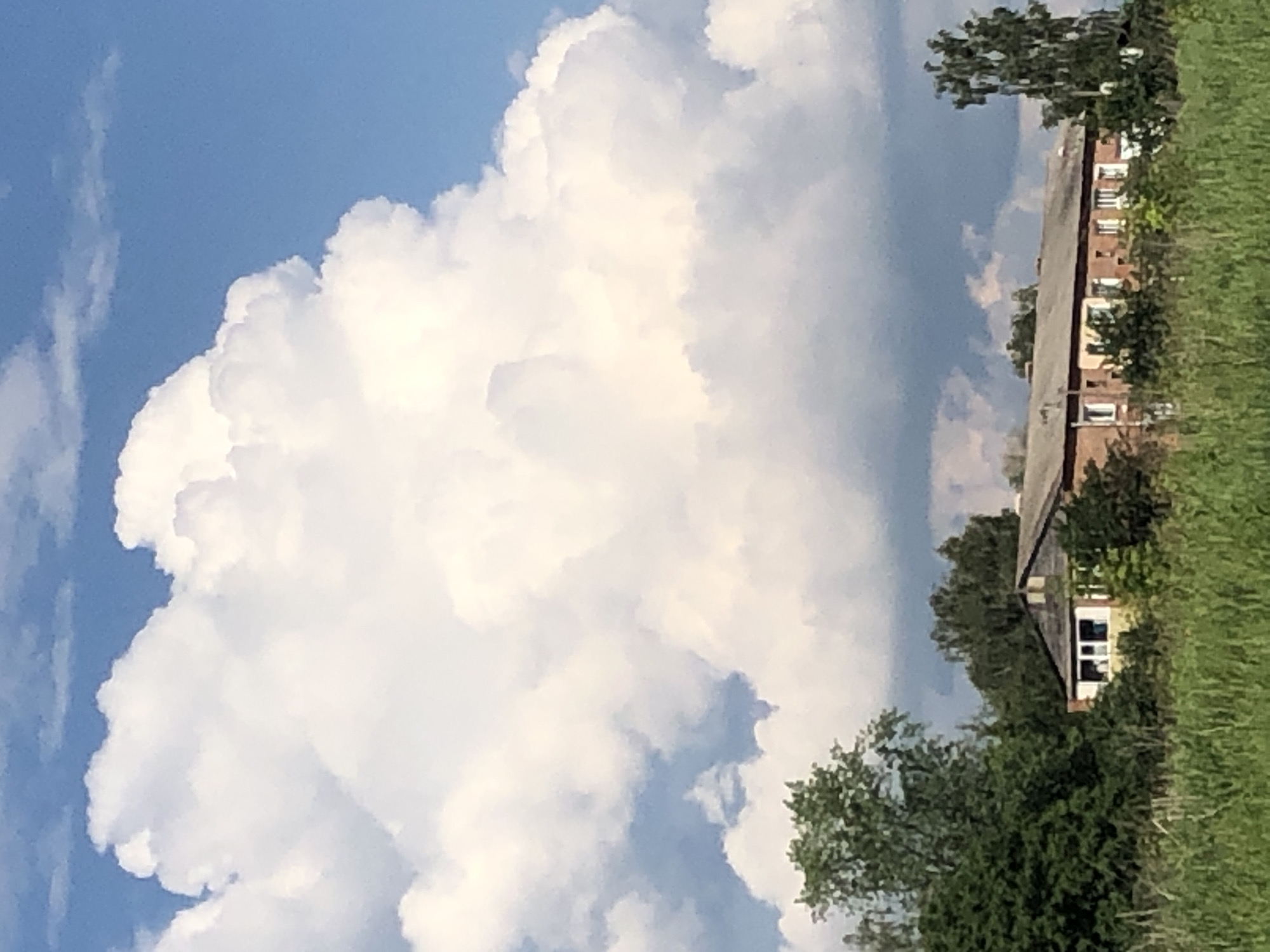The image captures a building, possibly an apartment block, nestled amongst trees and tall grass. The building is constructed of red brick with white-trimmed windows and a grey, pitched roof. There is a smaller, lighter-colored section attached to the left side of the main structure, also featuring white-trimmed windows. A large, dramatic cumulus cloud formation dominates the sky, casting a shadow over the scene and contrasting with the bright blue sky. The foreground is filled with lush green grass and foliage, suggesting a rural or semi-rural setting. The specific location of Khmilnytsia, Ukraine is indicated for this scene. No people are visible in the image.