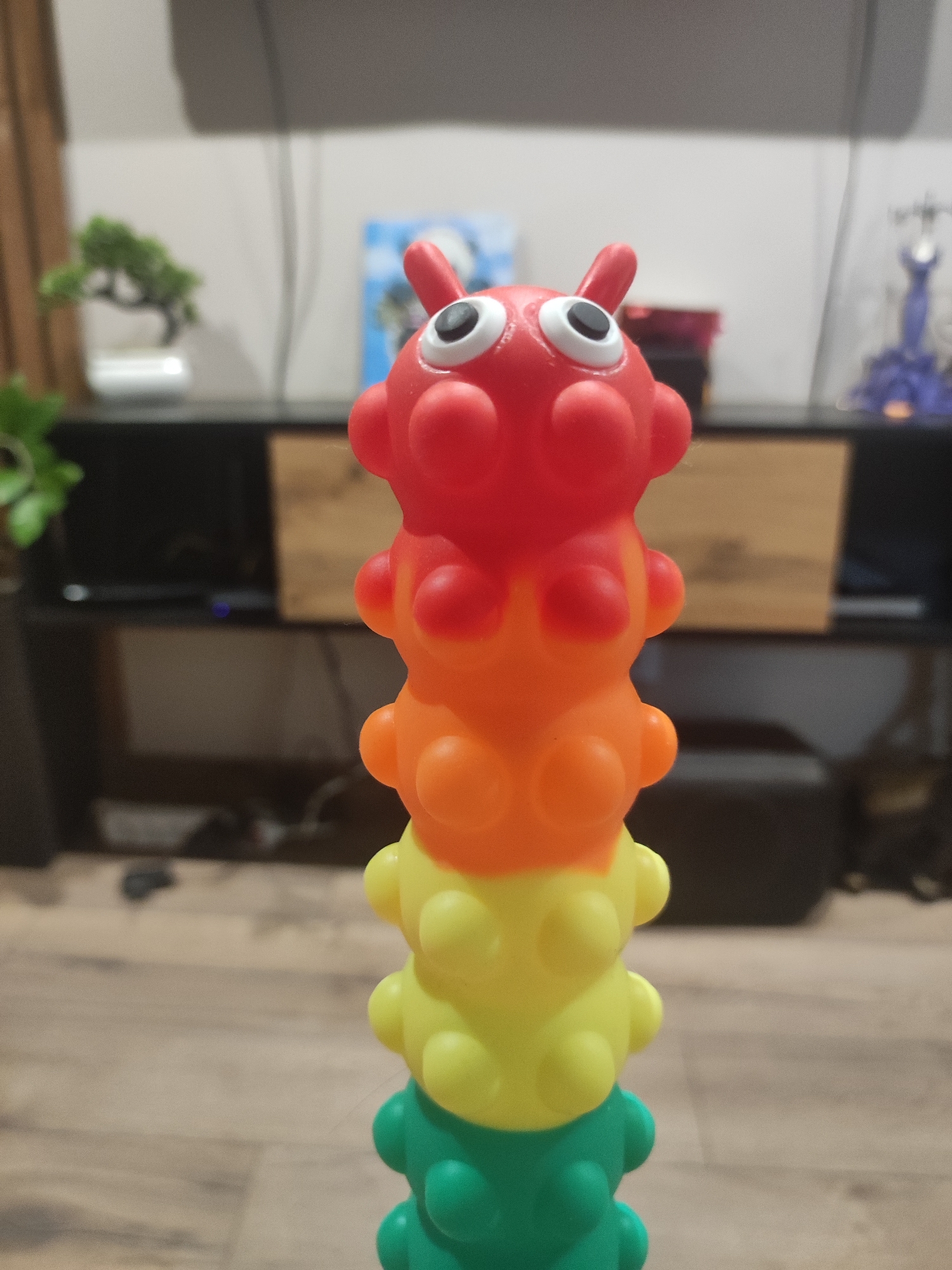 A close-up shot depicts a multi-colored toy caterpillar with large eyes, stacked in segments of red, orange, yellow, and green. The toy is positioned in the foreground, with a wooden floor beneath it. In the blurred background, there is a black entertainment center with various items on top, including plants, a small statue, and other decorative objects. The scene is indoors, and while the specific location in Votkinsk, Russia, is not discernible from the image alone, it can be inferred that it is a residential setting. There are no people visible in the shot. The focus is on the toy caterpillar.