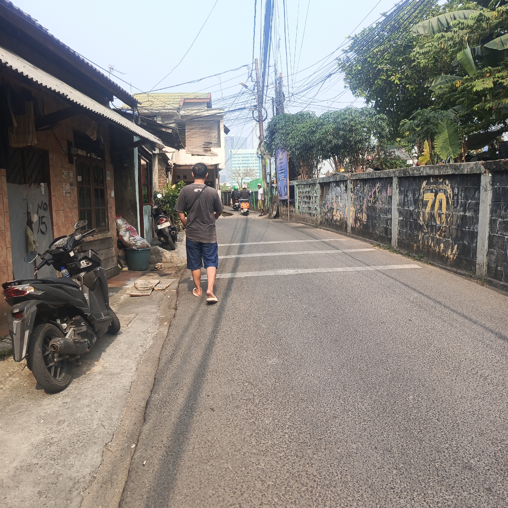 A man wearing a dark t-shirt and blue shorts walks away from the camera on a paved road in a city. He is wearing flip-flops. To his left, a black motorcycle is parked on a concrete sidewalk next to a building with a corrugated metal roof. The building has windows and doors, and graffiti or markings, including "No 15," are visible. Further down the sidewalk, there is a green trash bin with a large bag of trash next to it, and another motorcycle is parked behind it. Across the street, a concrete wall covered in graffiti runs along the side of the road. Trees and utility poles with numerous electrical wires are present. In the background, a modern building with a light-colored facade is visible, along with more trees and vegetation. The sky is clear and bright. The scene appears to be a street in a mixed urban area with residential and commercial elements.