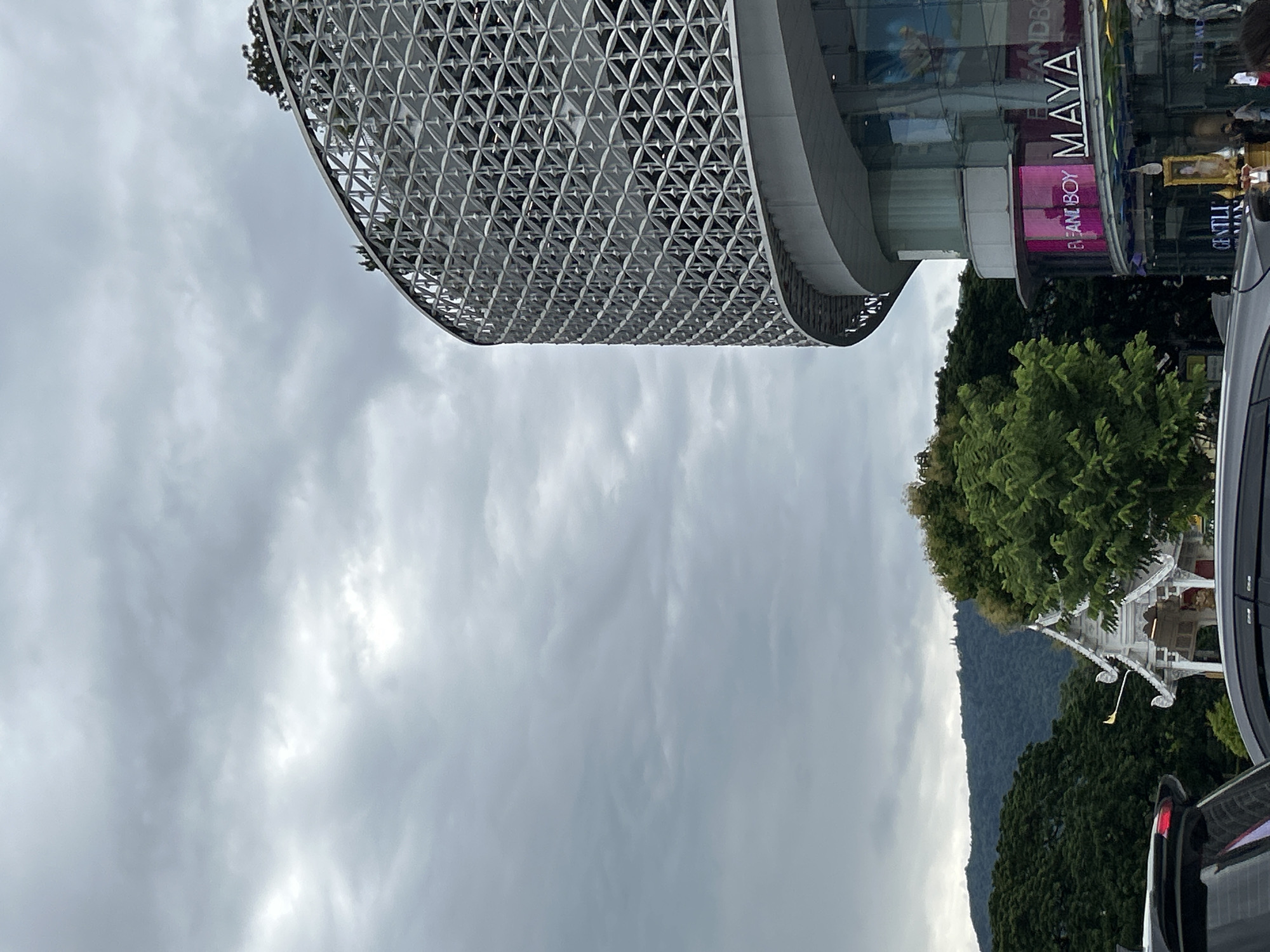 The image shows a modern building with a patterned facade and glass windows. A sign on the building reads "MAYA", suggesting it is a mall or commercial establishment. There are trees and a mountain in the background, indicating an outdoor setting, possibly in a city with natural surroundings. The sky is overcast, suggesting cloudy weather. The scene appears to be a daytime view, but the light is diffused due to the clouds. There are some vehicles visible, indicating it's a public area. The overall impression is one of an urban environment integrated with nature. The text "ELEPHANT BOY" is also visible on a pink sign.
