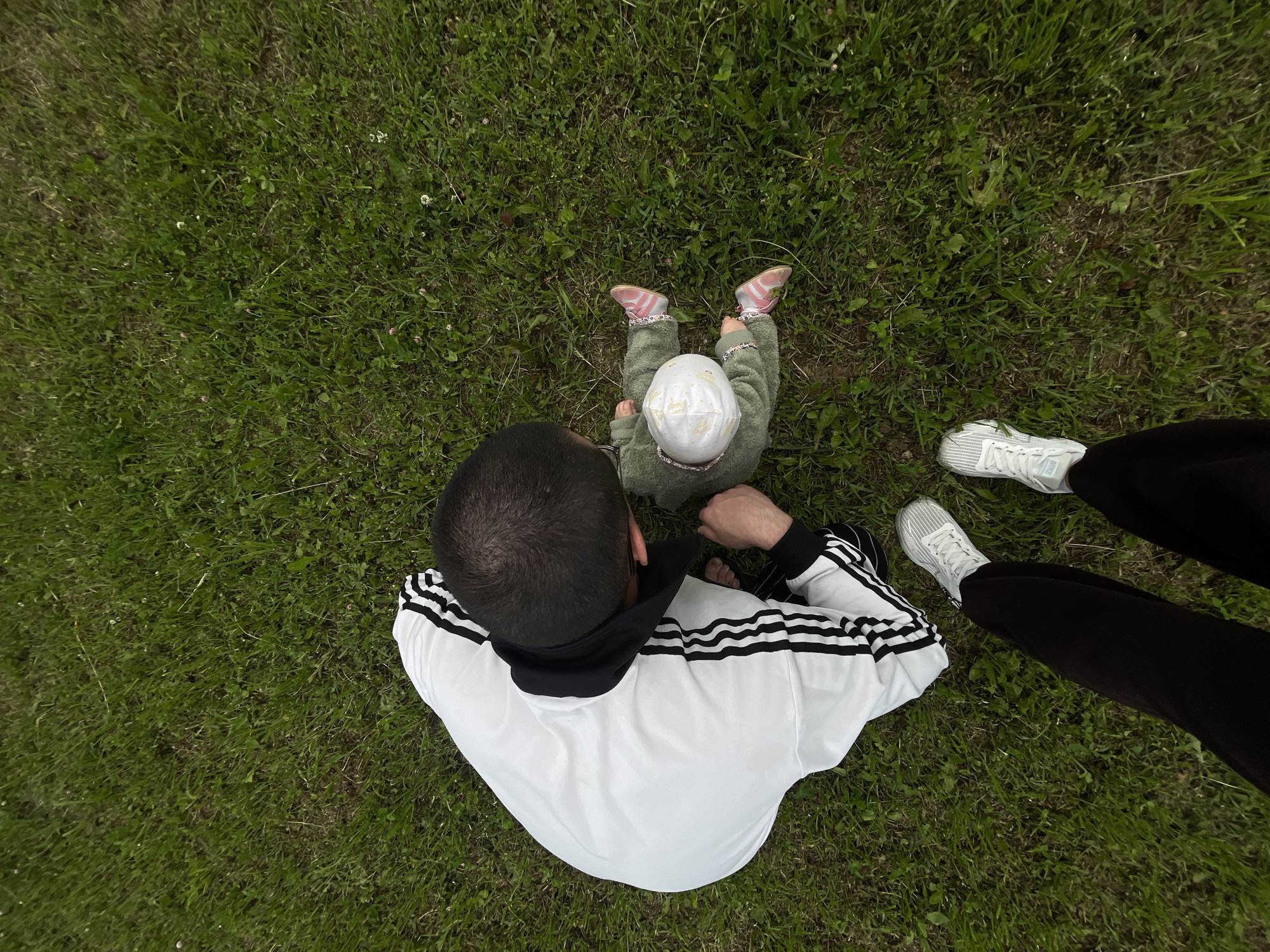 A person in a white tracksuit with black stripes kneels on a grassy area, looking down at a baby sitting on the grass. The baby is wearing a light green outfit and a white hat. The person's feet are visible in the foreground, wearing dark pants and white athletic shoes. The environment is a field of green grass, suggesting an outdoor setting. There is no indication of the city of Подшивалово, Russia.