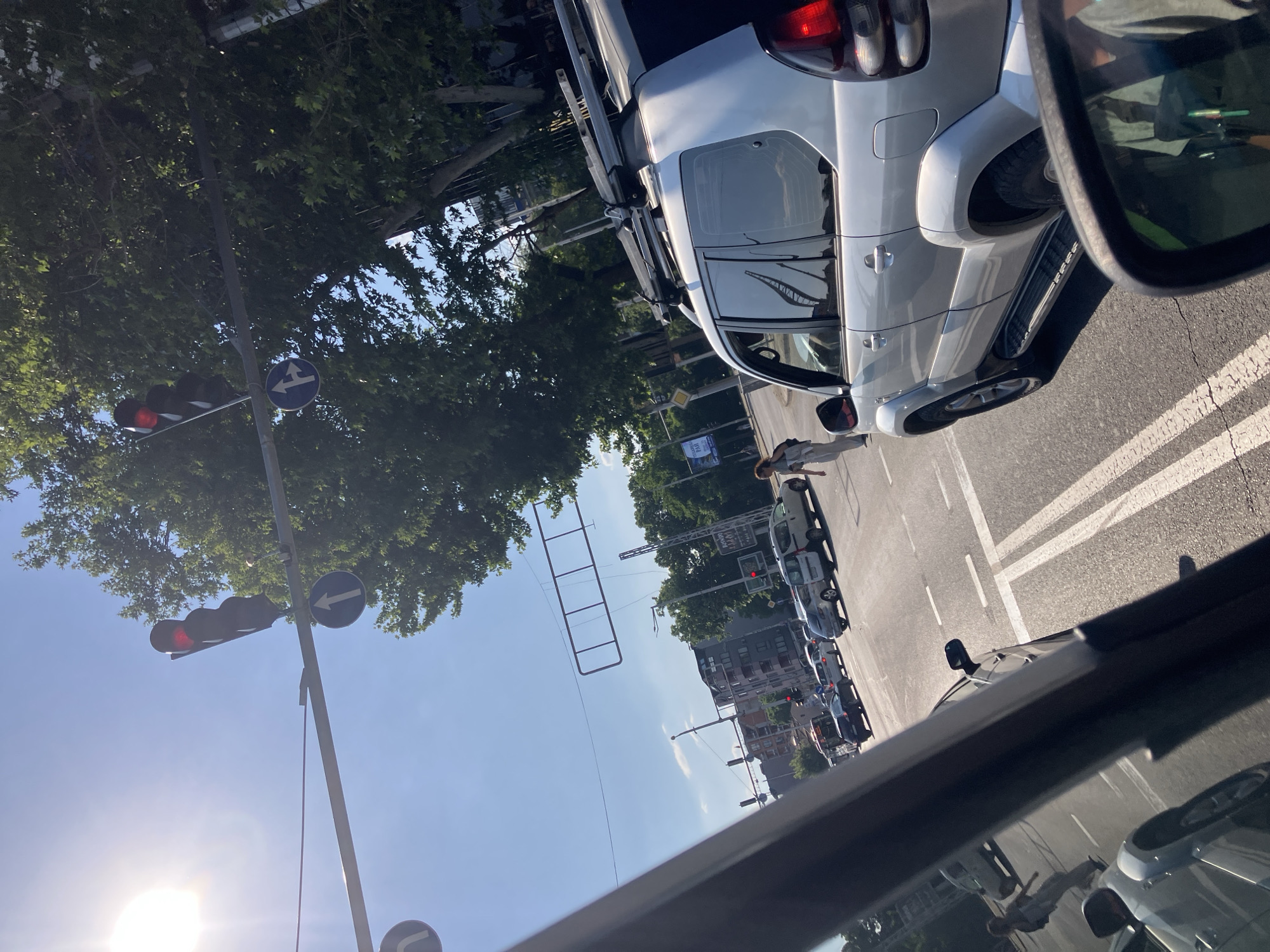 The image shows a street scene in Plovdiv, Bulgaria. The view is from inside a car, looking out onto a street with traffic. A silver SUV with roof racks is adjacent to the viewer. There are several other cars parked along the street. A pedestrian is crossing the street. Overhead traffic signals are visible, showing red lights. Street signs are present. The sky is blue with some scattered trees. Buildings are visible in the background.